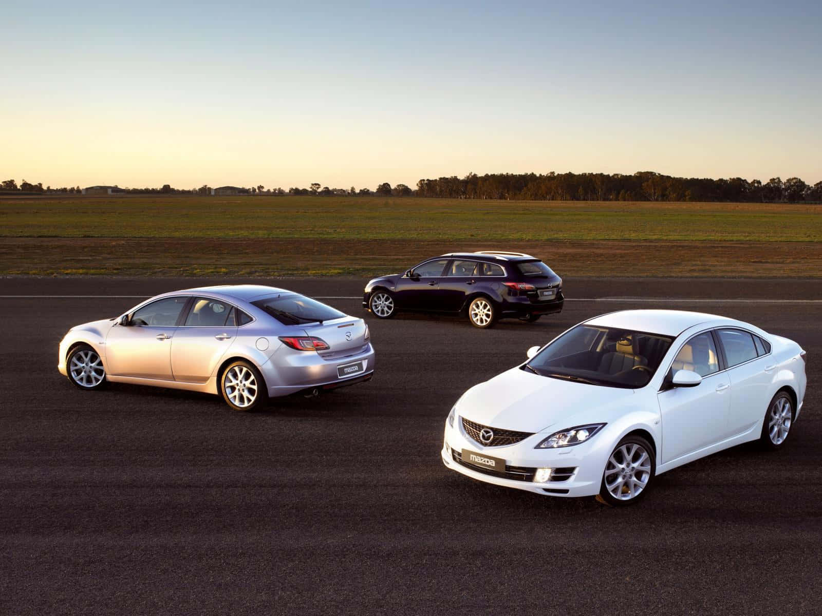 Mazda 6 桌布 壁紙 — 三輛萬事得房車（銀色、黑色、白色）停泊在瀝青路上，金色夕陽照亮開闊田野。
