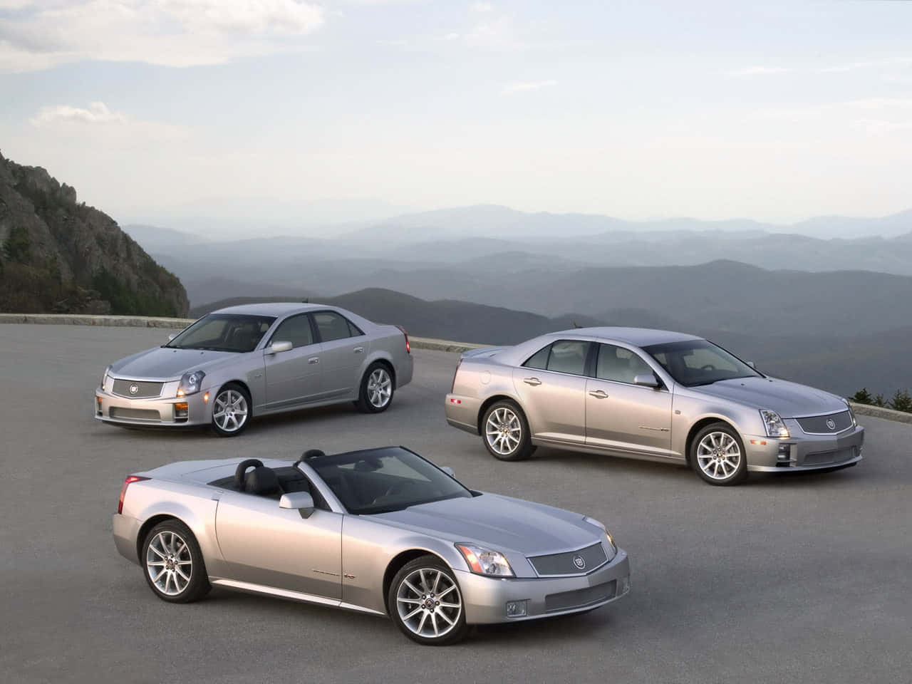 Cadillac Xlr 桌布 壁紙 — 三輛銀色Cadillac汽車停在山頂觀景點，層層山巒在柔和日光下逐漸消失在霧氣瀰漫的遠方。