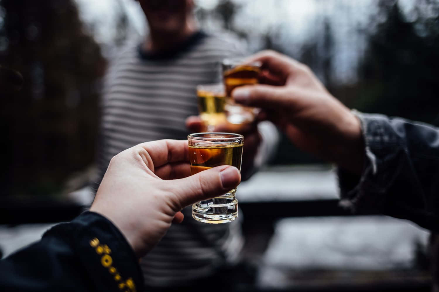 醉咗 桌布 壁紙 — 三隻手在模糊的戶外環境中碰杯，杯中裝滿琥珀色液體。