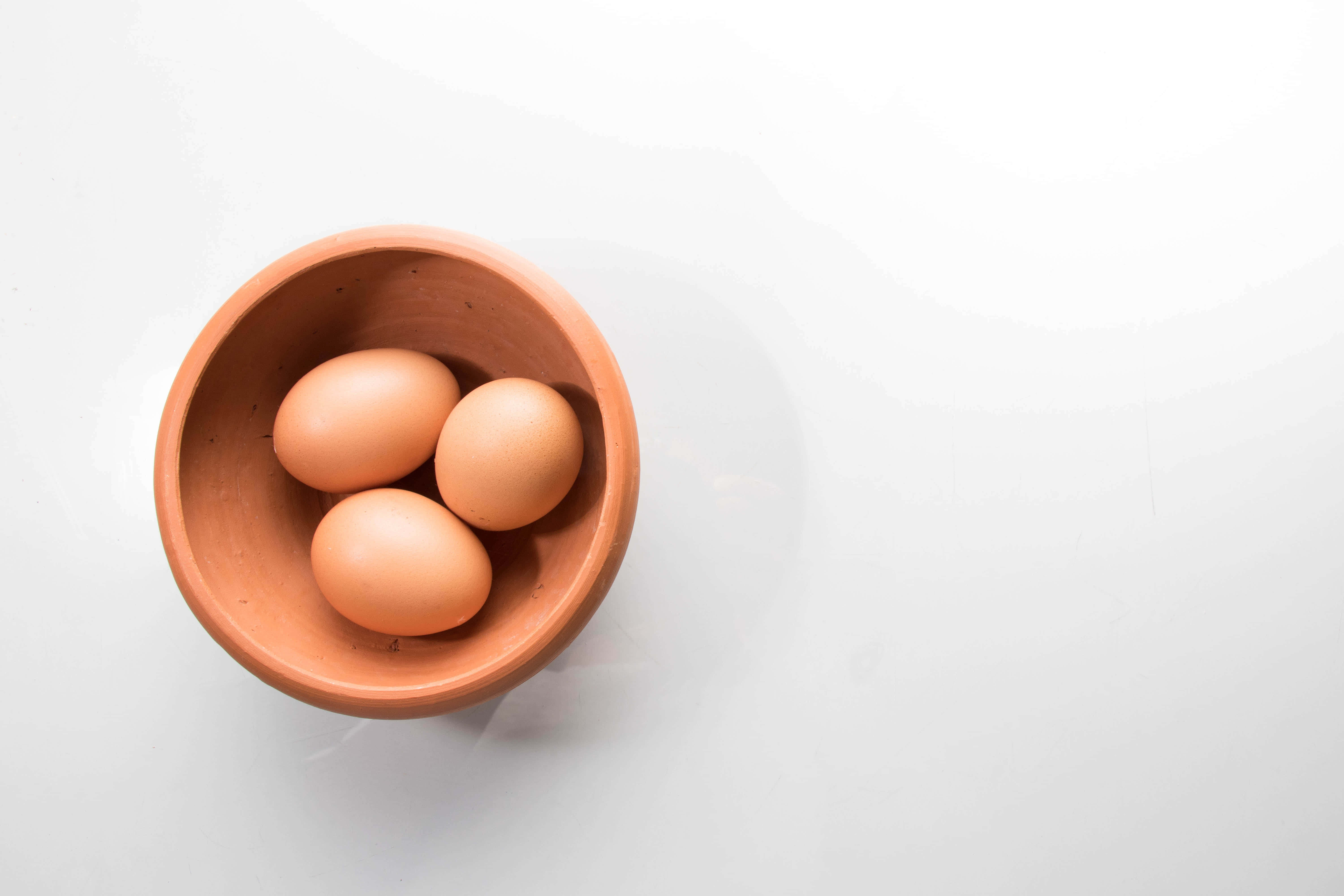 Brown Eggs 桌布 壁紙 — 三隻棕色雞蛋放在一個赤陶碗中，置於亮白色表面上，從正上方俯瞰。
