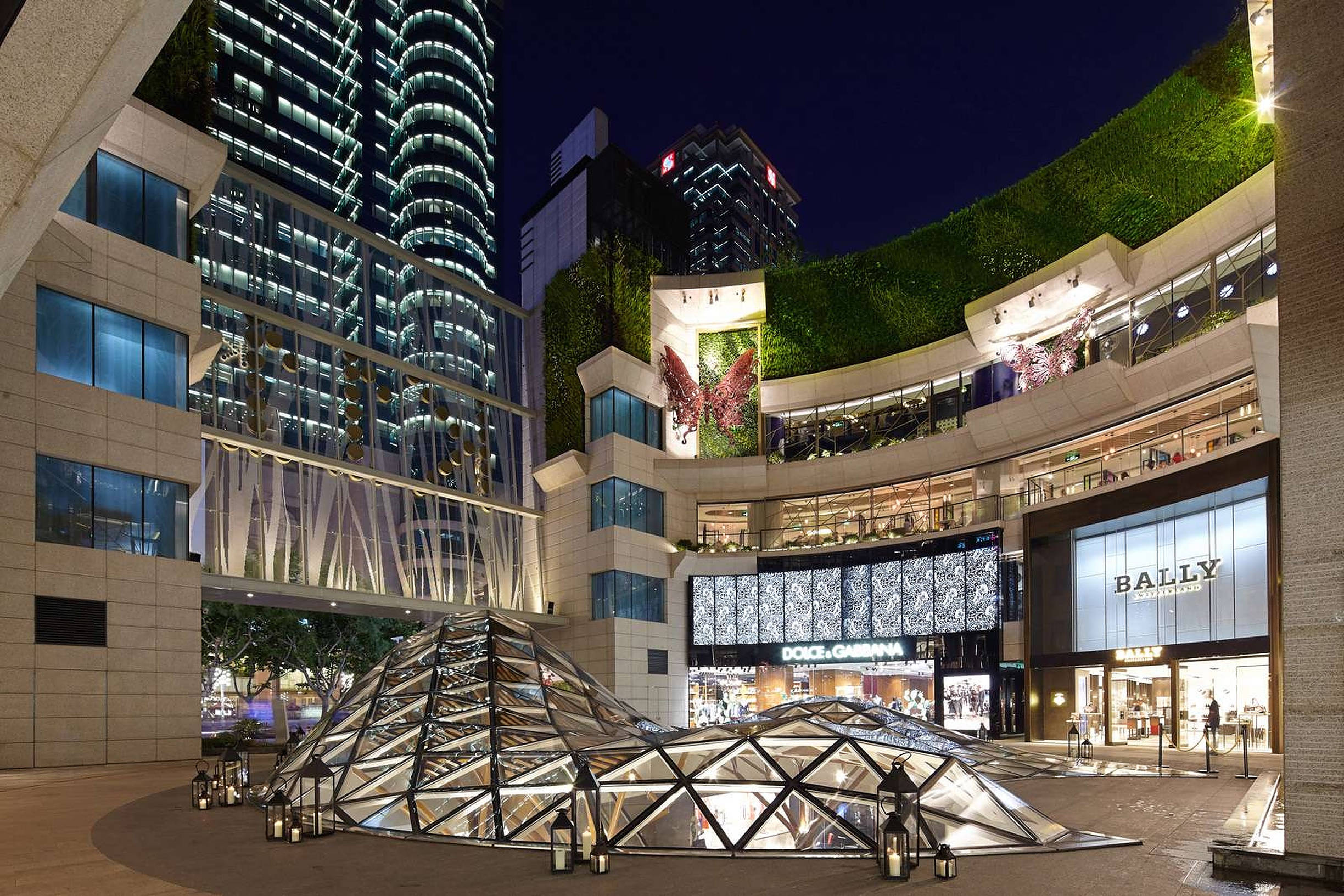 上海K11購物廣場 壁紙 — 一個耀眼的夜間城市廣場，特色是玻璃圓頂結構，周圍環繞著燈火通明的高層建築和茂密的綠色牆壁。