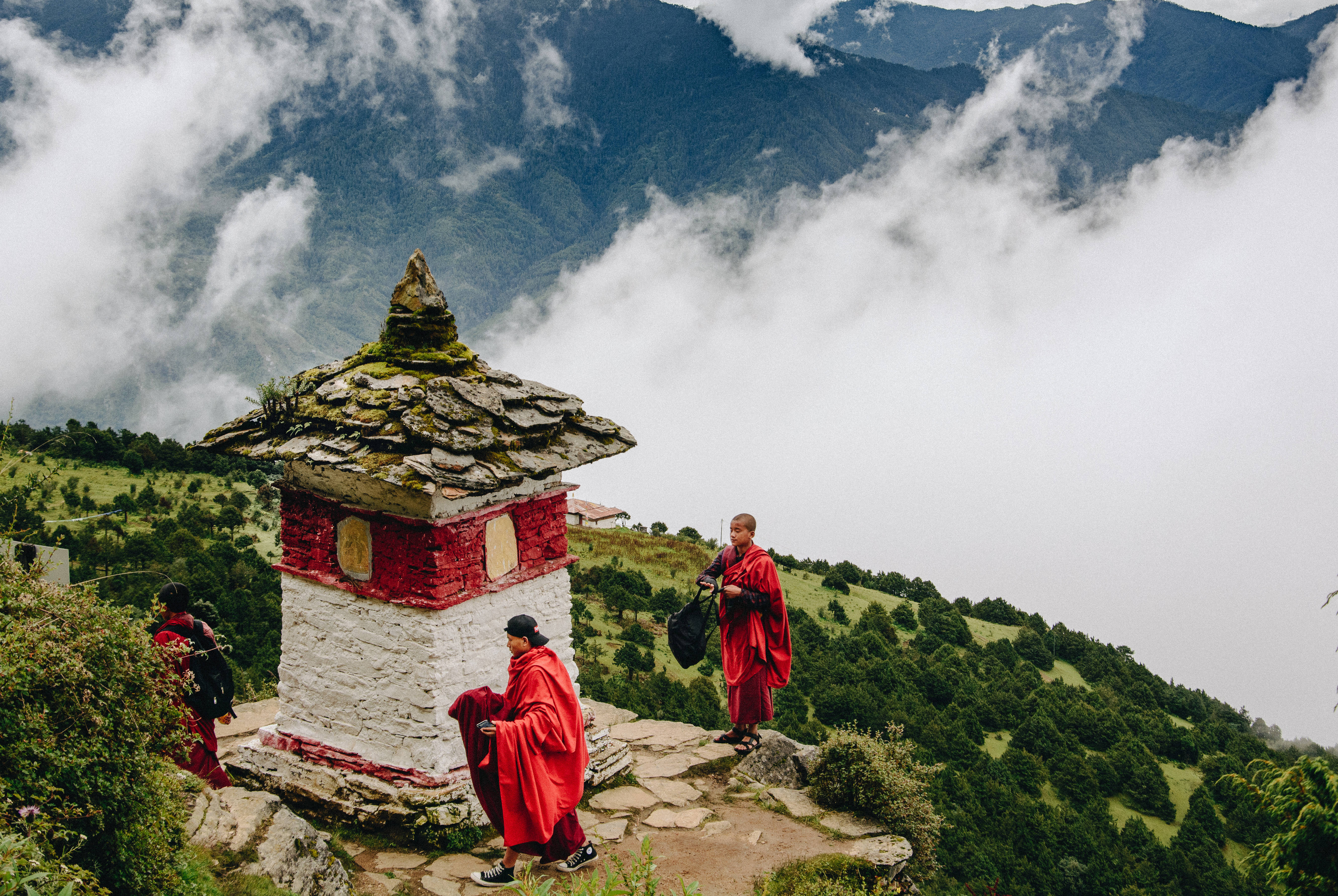 不丹圖吉德拉格寺 壁紙 — 身穿鮮紅色僧袍的僧侶在雲霧繚繞的山脊上，沿著一座石砌佛塔行走，俯瞰雲海覆蓋的山谷。