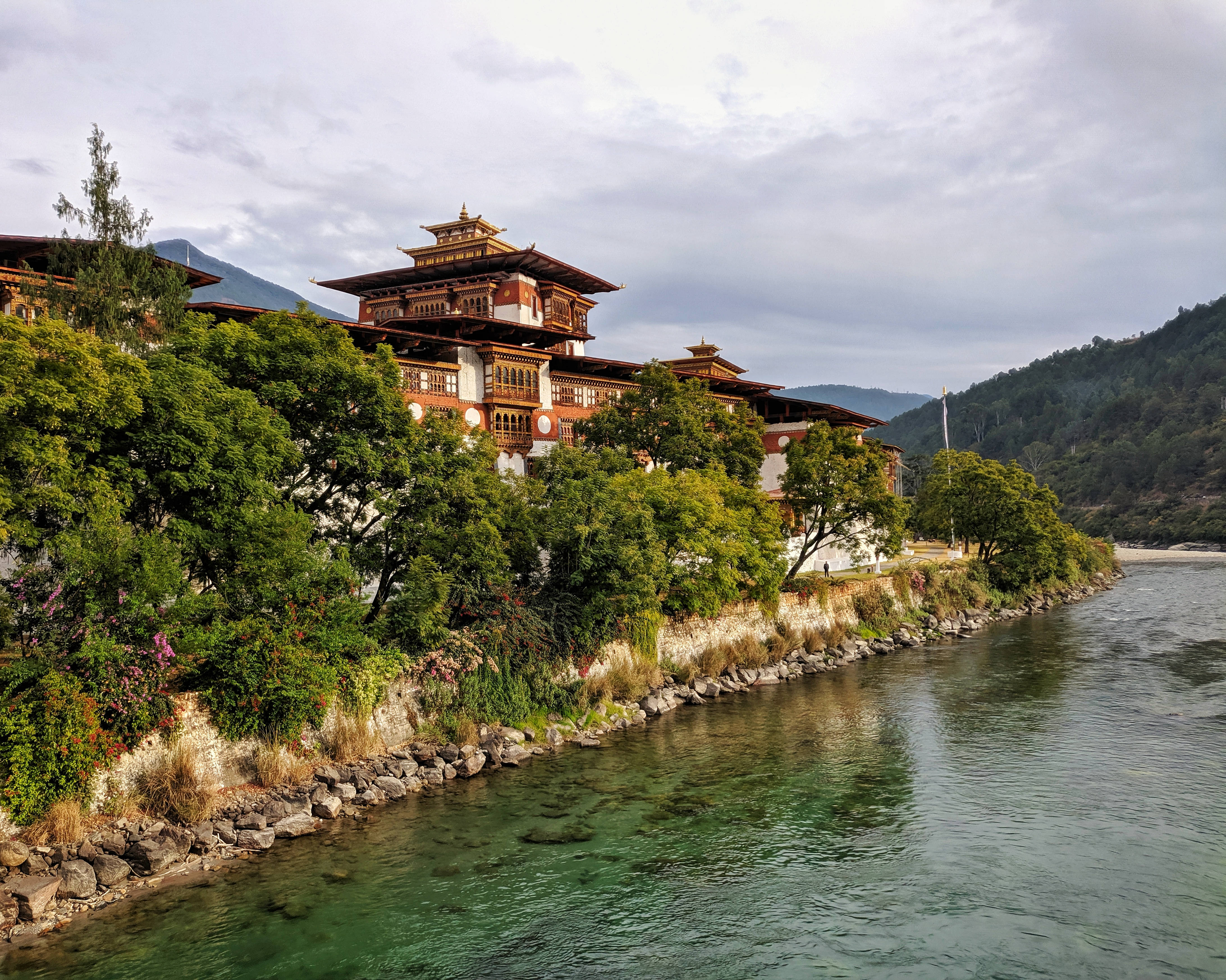 不丹彭措拉宮 壁紙 — 傳統不丹寺廟，層疊屋頂，坐落於平靜的河邊，兩旁綠樹成蔭，天空多雲。