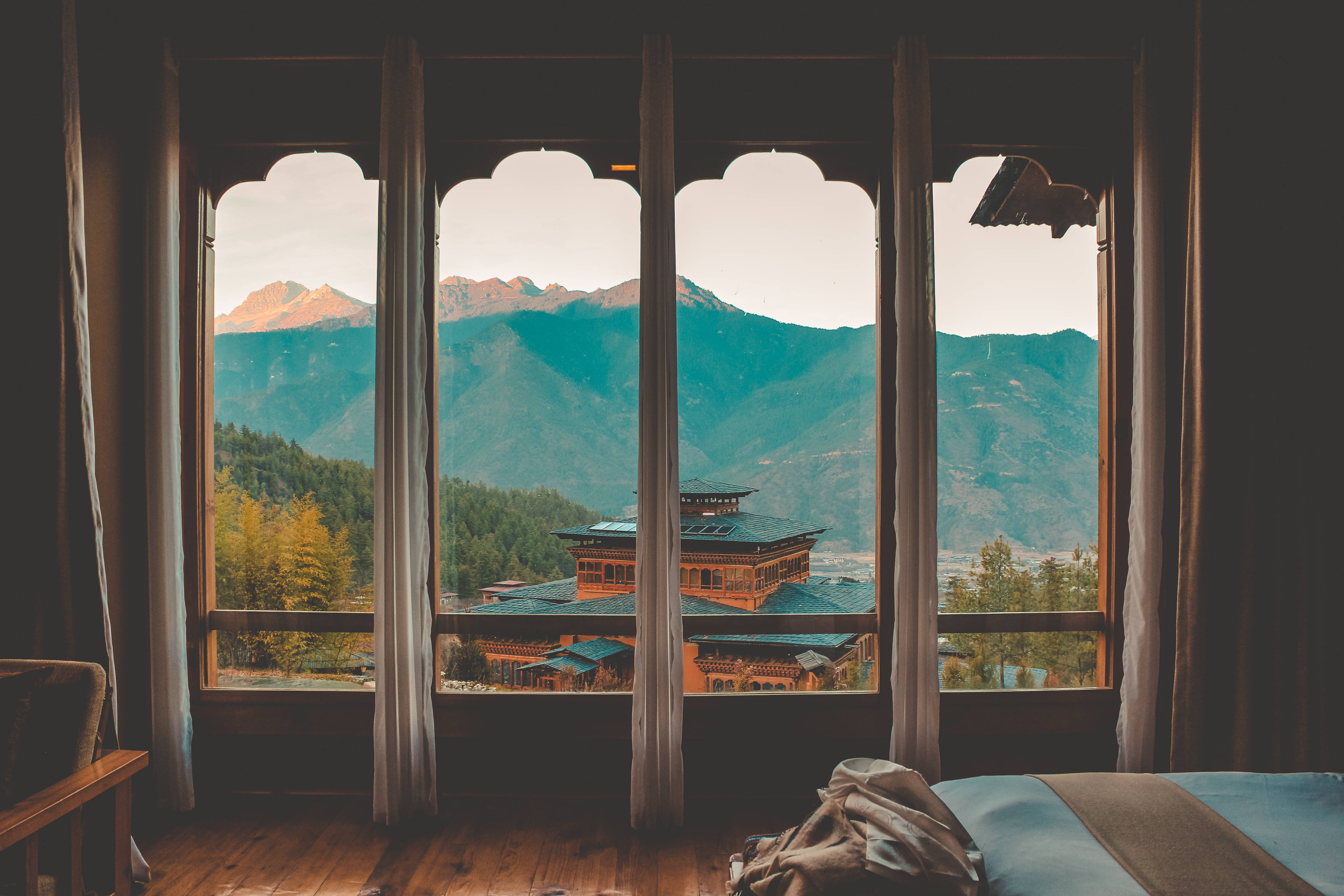 不丹普姆奇哈旅館景觀 壁紙 — 舒適的臥室，木地板和窗簾框住了日出時分鬱鬱蔥蔥的綠色山巒中傳統不丹寺廟的景色。