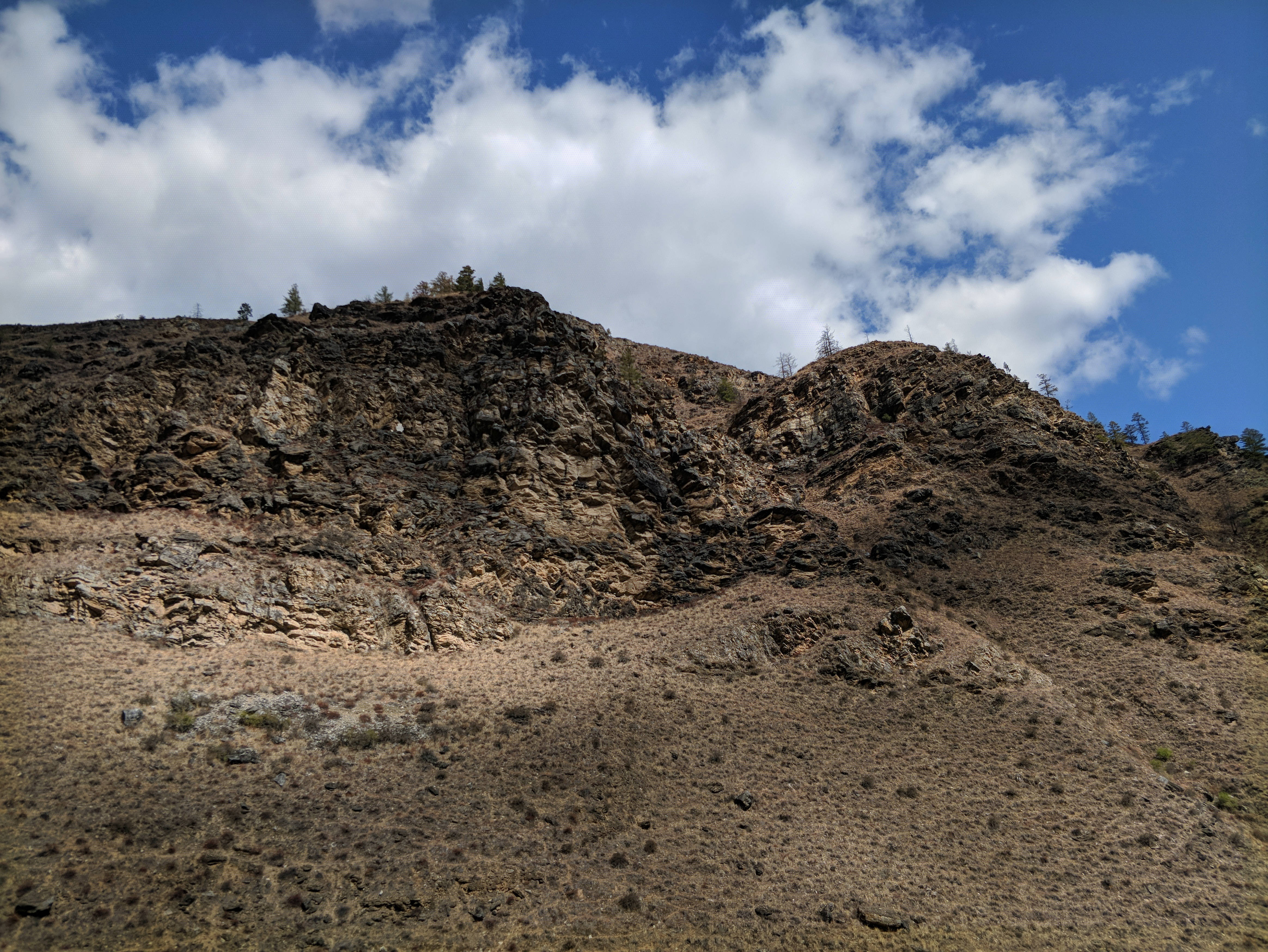 不丹楚克哈山 壁紙 — 崎嶇的火山山坡，有深色岩石露頭和稀疏的乾燥植被，在明亮的藍天下有蓬鬆的白雲。