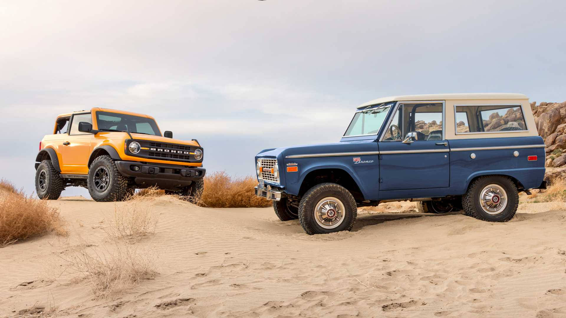不同款式的福特 Bronco 壁紙 — 兩架復古Bronco，一架橙色一架藍色，停泊喺沙丘上，背景係岩石山丘，天空多雲。