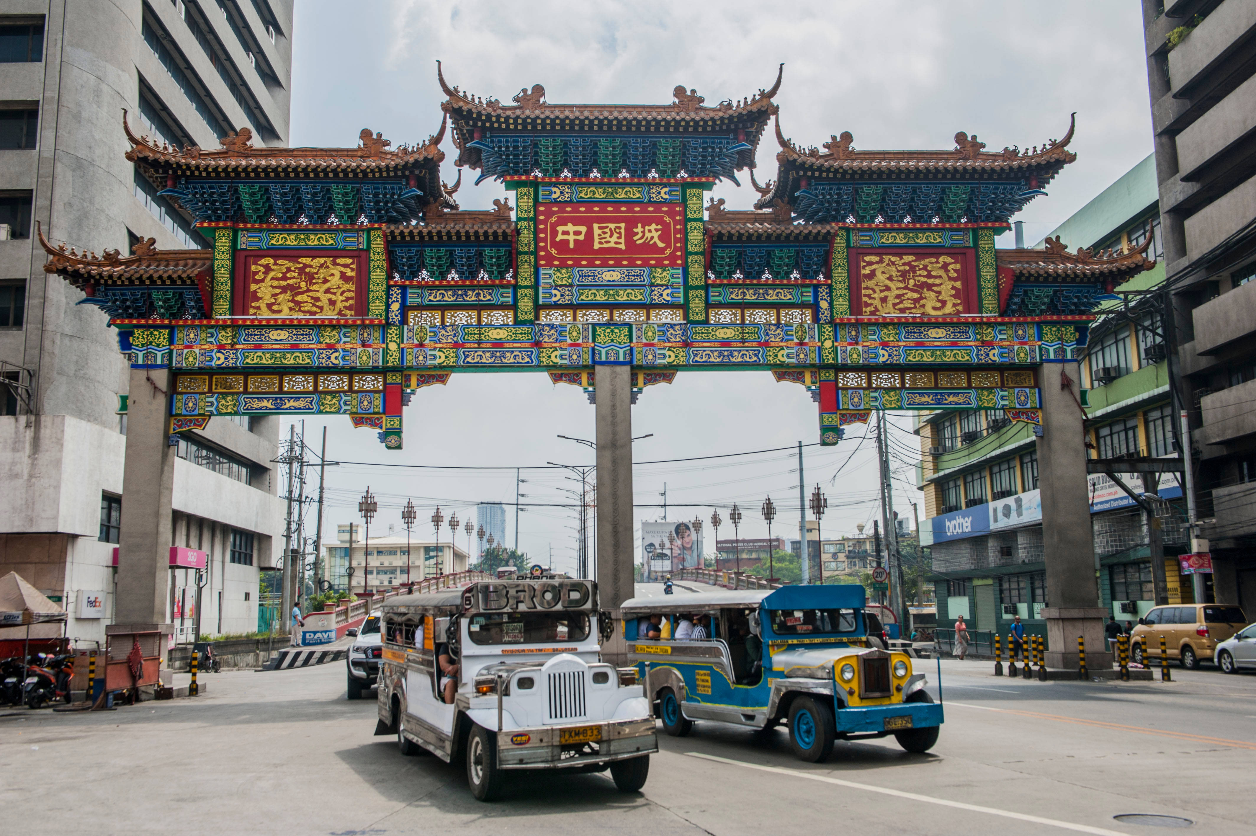 世界最古老唐人街馬尼拉 壁紙 — 色彩繽紛的傳統中式牌坊，帶有精緻的紅、金、藍色細節，矗立在繁忙都市街道上色彩繽紛的吉普尼車上方。