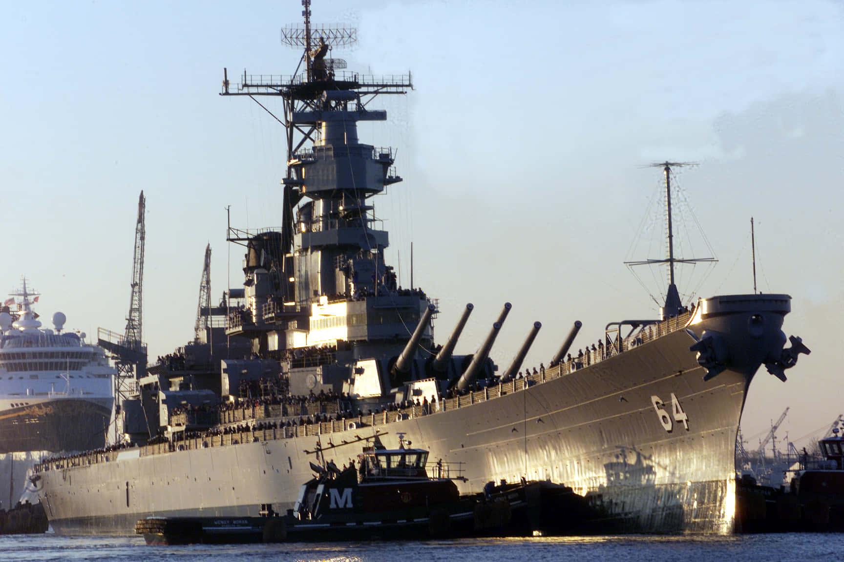 世界海軍傳統的象徵 壁紙 — 一艘巨大的灰色戰艦，編號64，帶有突出的炮塔，停泊在港口，附近有遊輪和拖船。