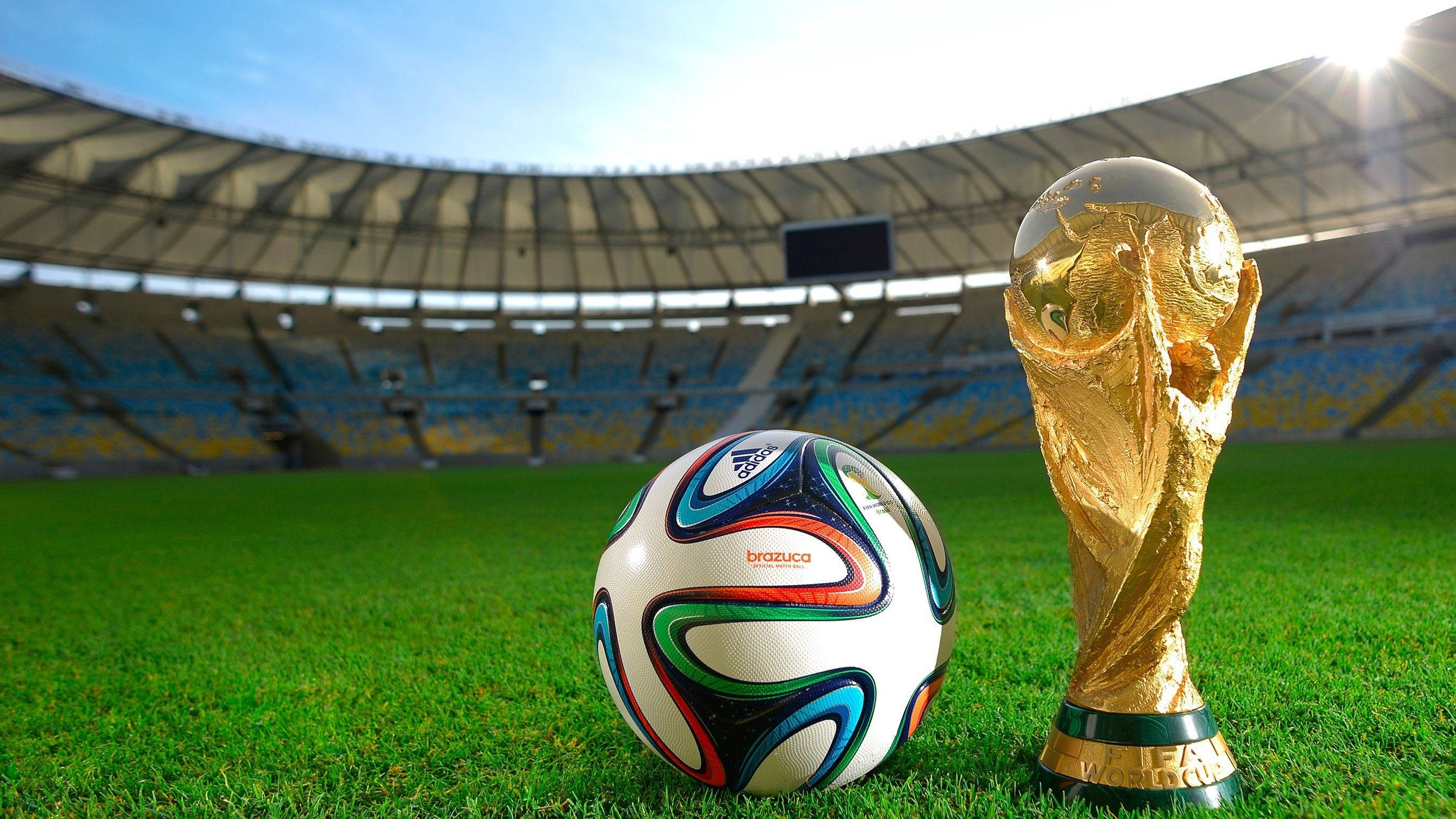 世界盃與足球 壁紙 — 閃閃發光的世界盃獎盃和色彩繽紛的 Brazuca 足球，在燦爛陽光下，安放在一個巨大空蕩蕩的體育館內生機勃勃的綠草地上。