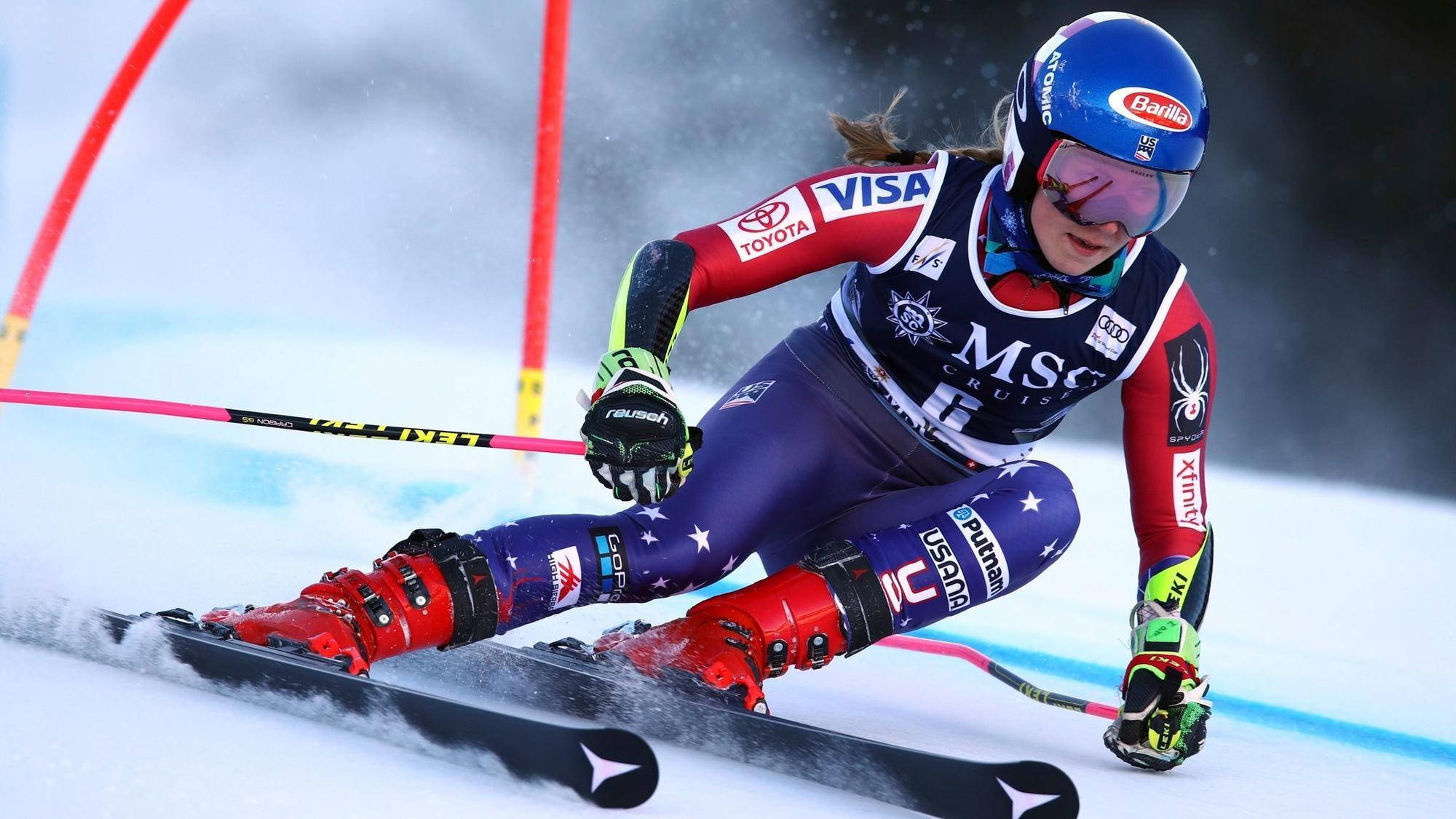 世界知名高山滑雪冠軍Mikaela Shiffrin下坡競技 壁紙 — 一位戴著藍色頭盔和愛國裝備的女性高山滑雪運動員在下坡比賽中專注地切過積雪。