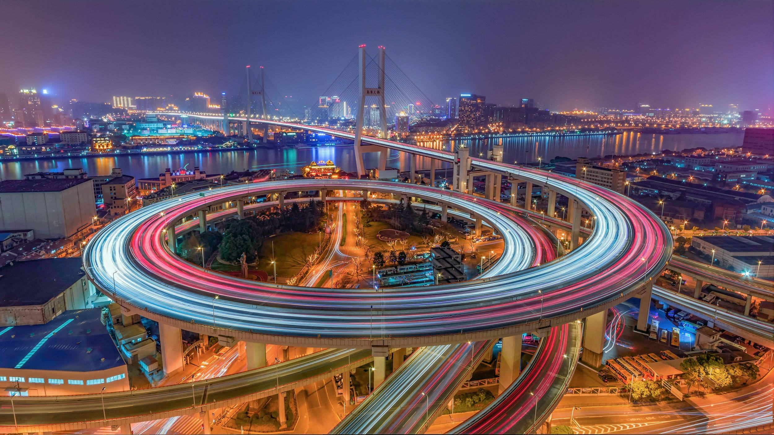 中國上海南浦大橋夜景 壁紙 — 令人眼花繚亂的夜間延時攝影，呈現螺旋形高速公路交匯處，車燈流光溢彩，照亮城市景觀和河流。