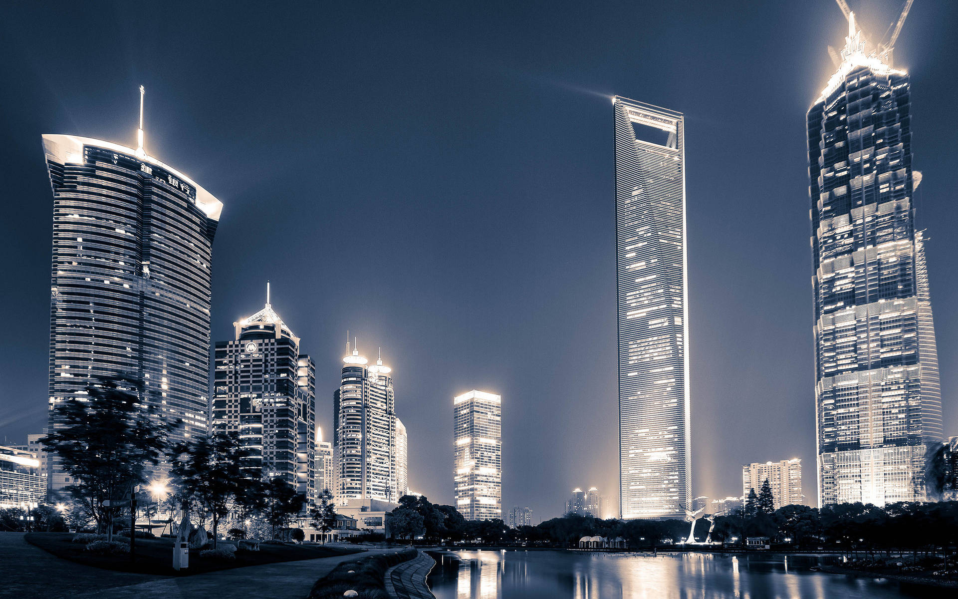 中國上海現代都市 壁紙 — 高聳的摩天大樓在深藍色的夜空中閃耀，它們的燈光倒映在下方平靜的海濱上。
