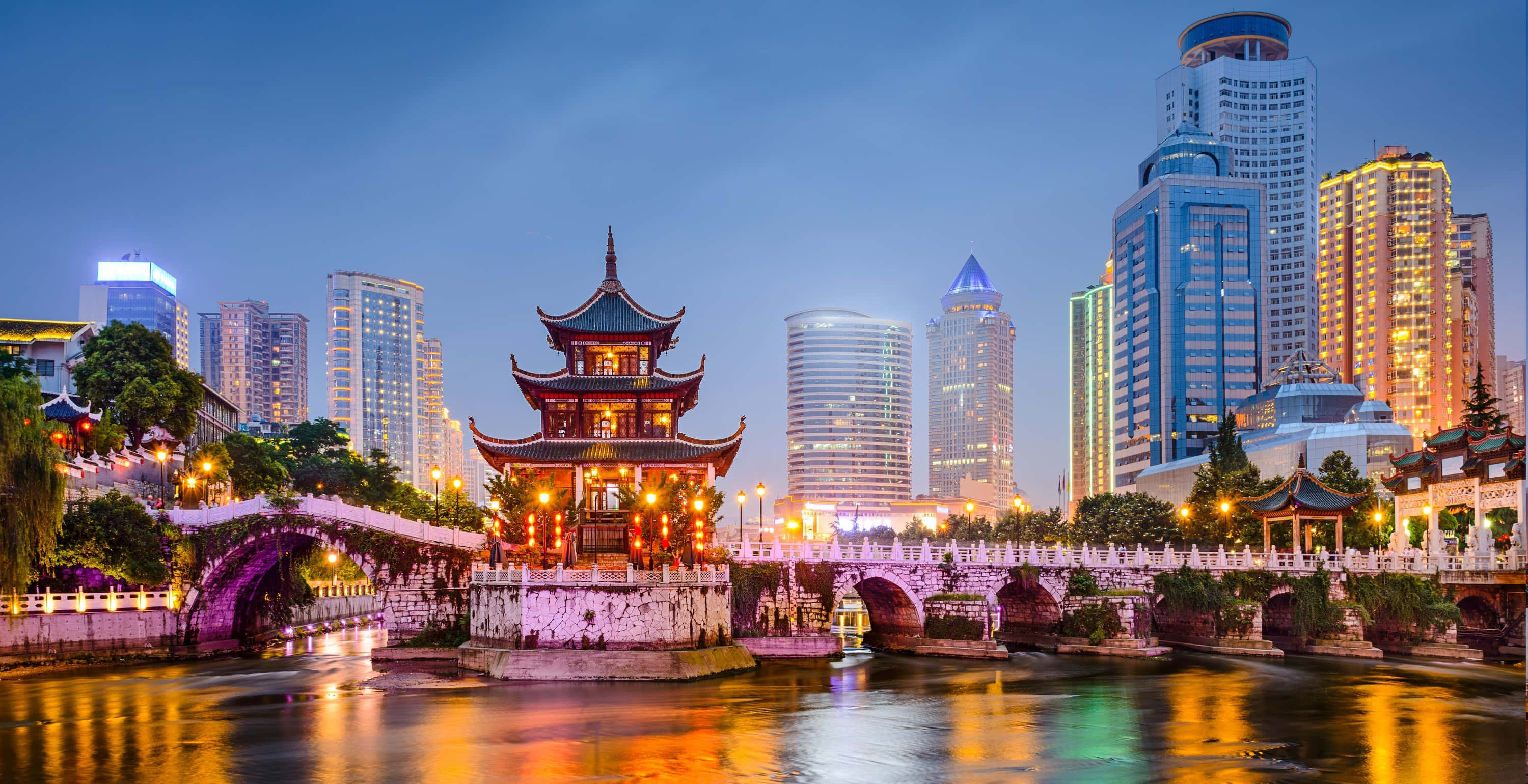中國之美 壁紙 — 一座傳統中式亭閣在黃昏的河上溫暖地發光，被水中倒映的現代摩天大樓所環繞。