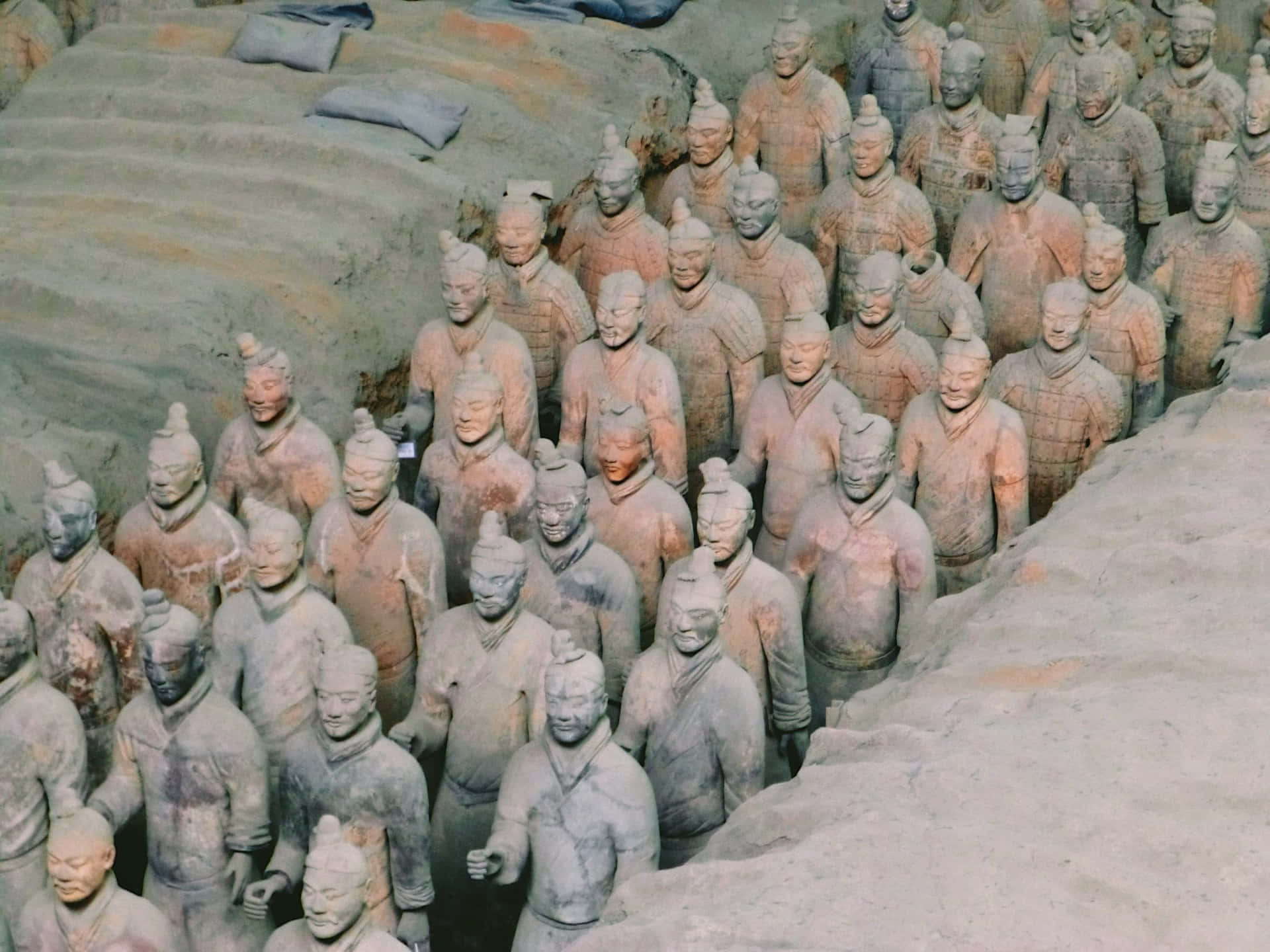 中國博物館石質陶俑雕塑 壁紙 — 一排排古老的兵馬俑以大地色調矗立在它們的發掘坑中，臉部細節帶有莊嚴的表情。