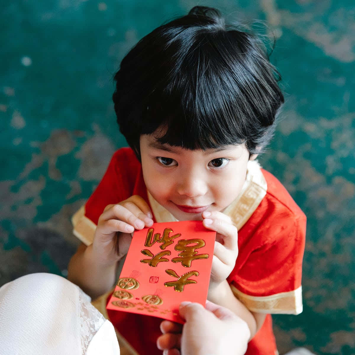 中國古水鄉 壁紙 — 一個穿著紅色傳統服裝的小孩拿著一個帶有金色字樣的紅色利是封，抬頭露出溫柔的微笑。