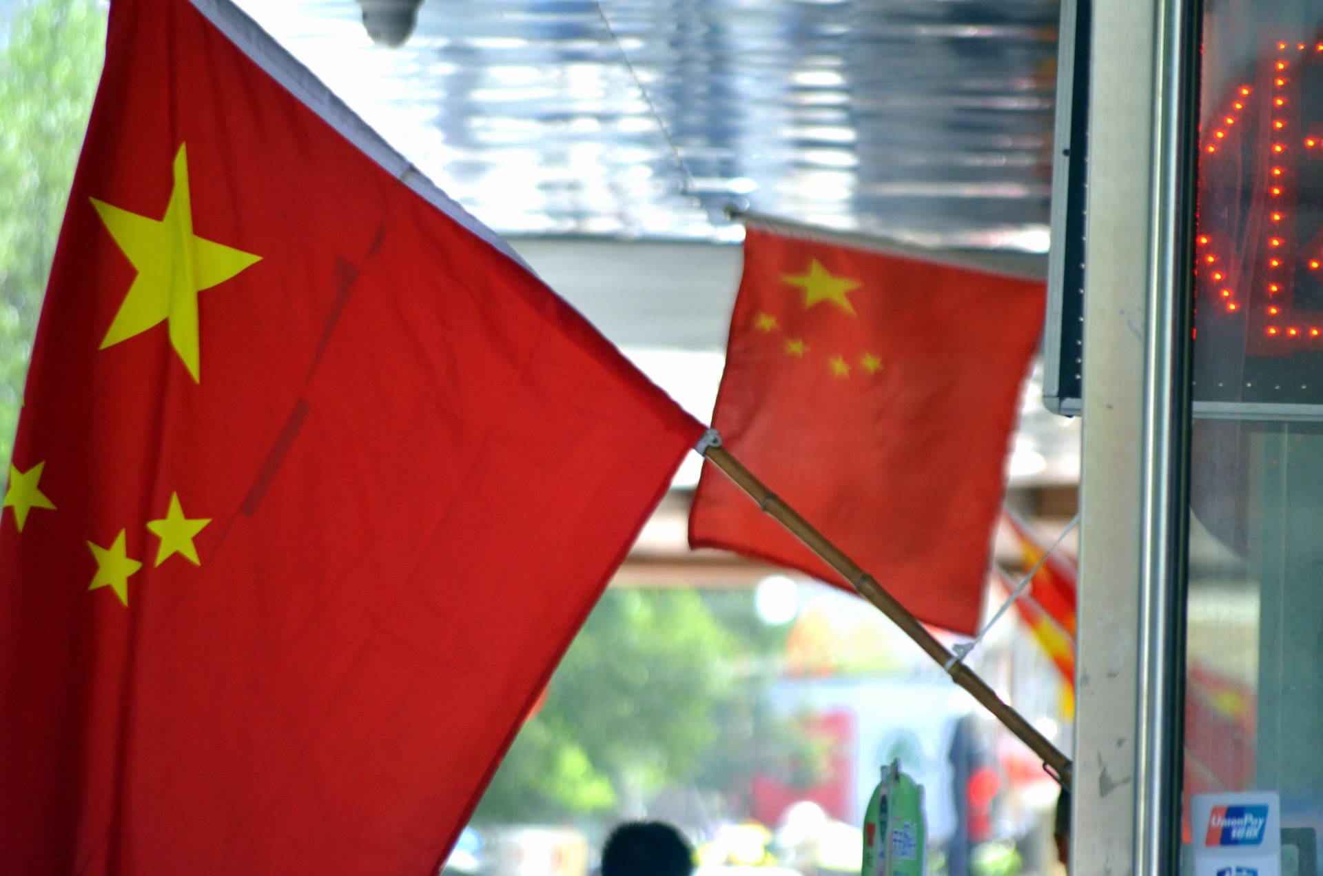 中國國旗商店 壁紙 — 兩面中國國旗掛在室內近窗位置，窗外有模糊的戶外綠植和發光招牌。