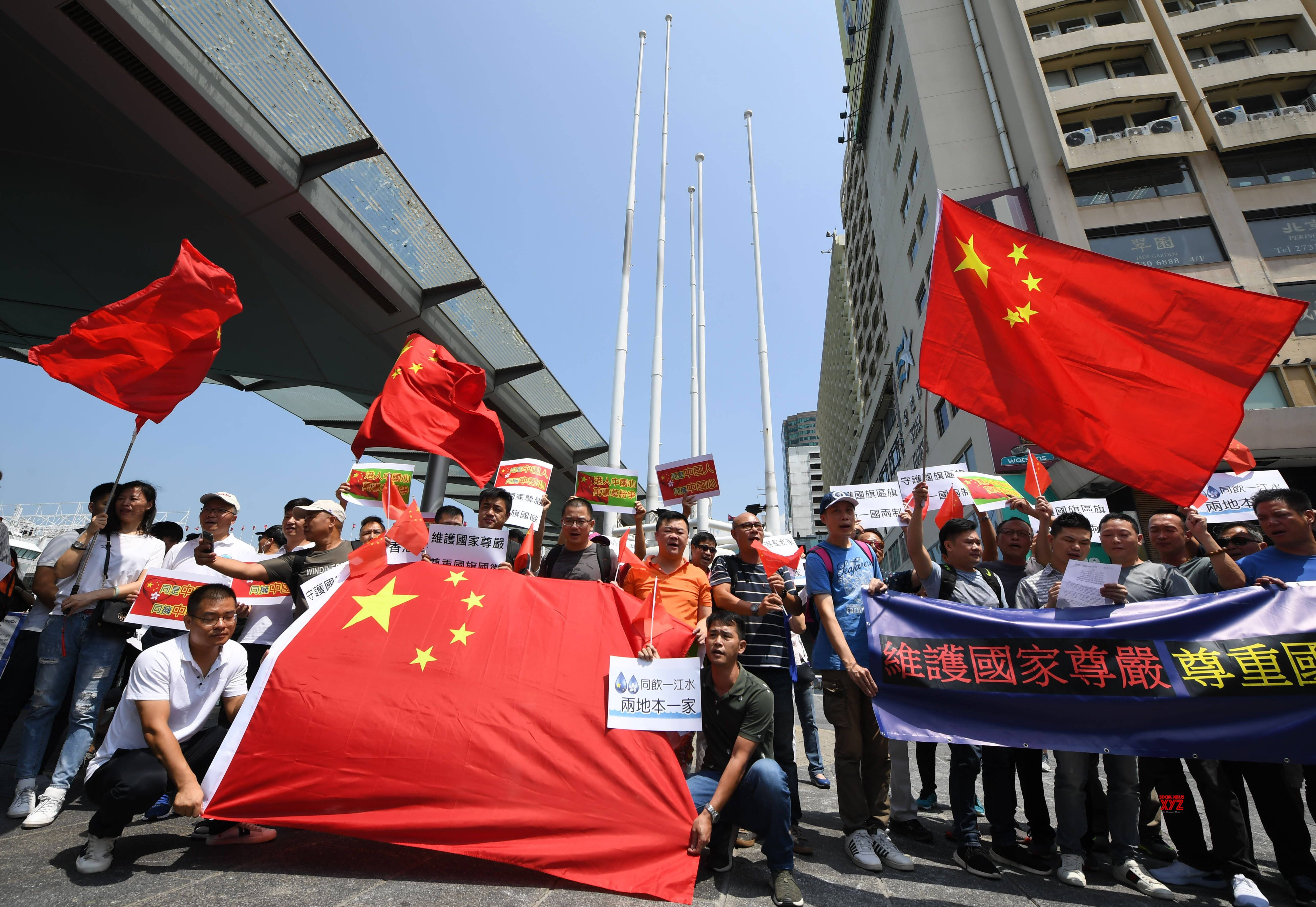中國國旗抗議 壁紙 — 一群人在陽光明媚的城市街道上，在現代建築之間手持大型紅色中國國旗和標語抗議。