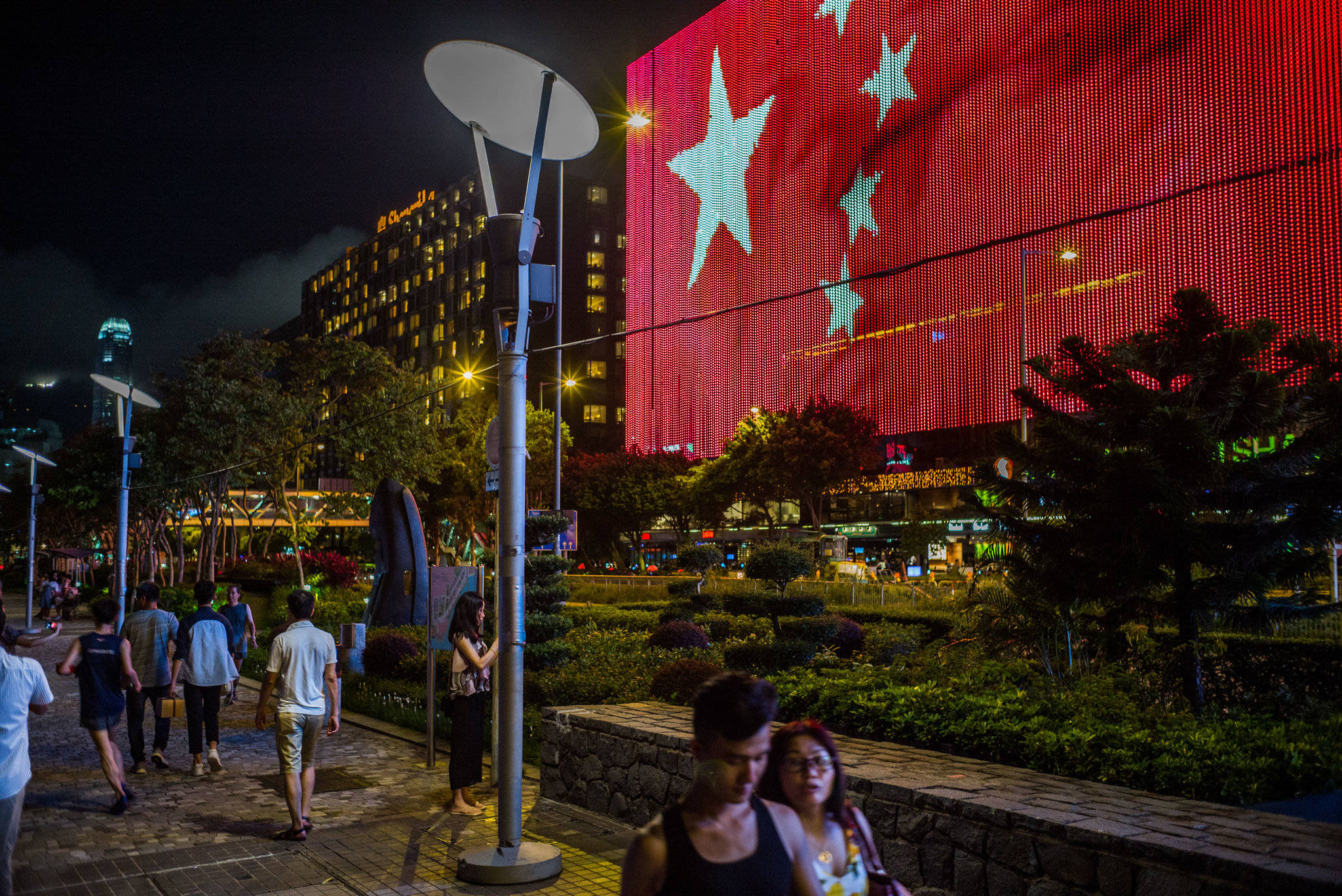 中國國旗螢幕 壁紙 — 充滿活力的夜間城市場景，行人走過建築物外牆上巨大的紅色旗幟燈光展示。