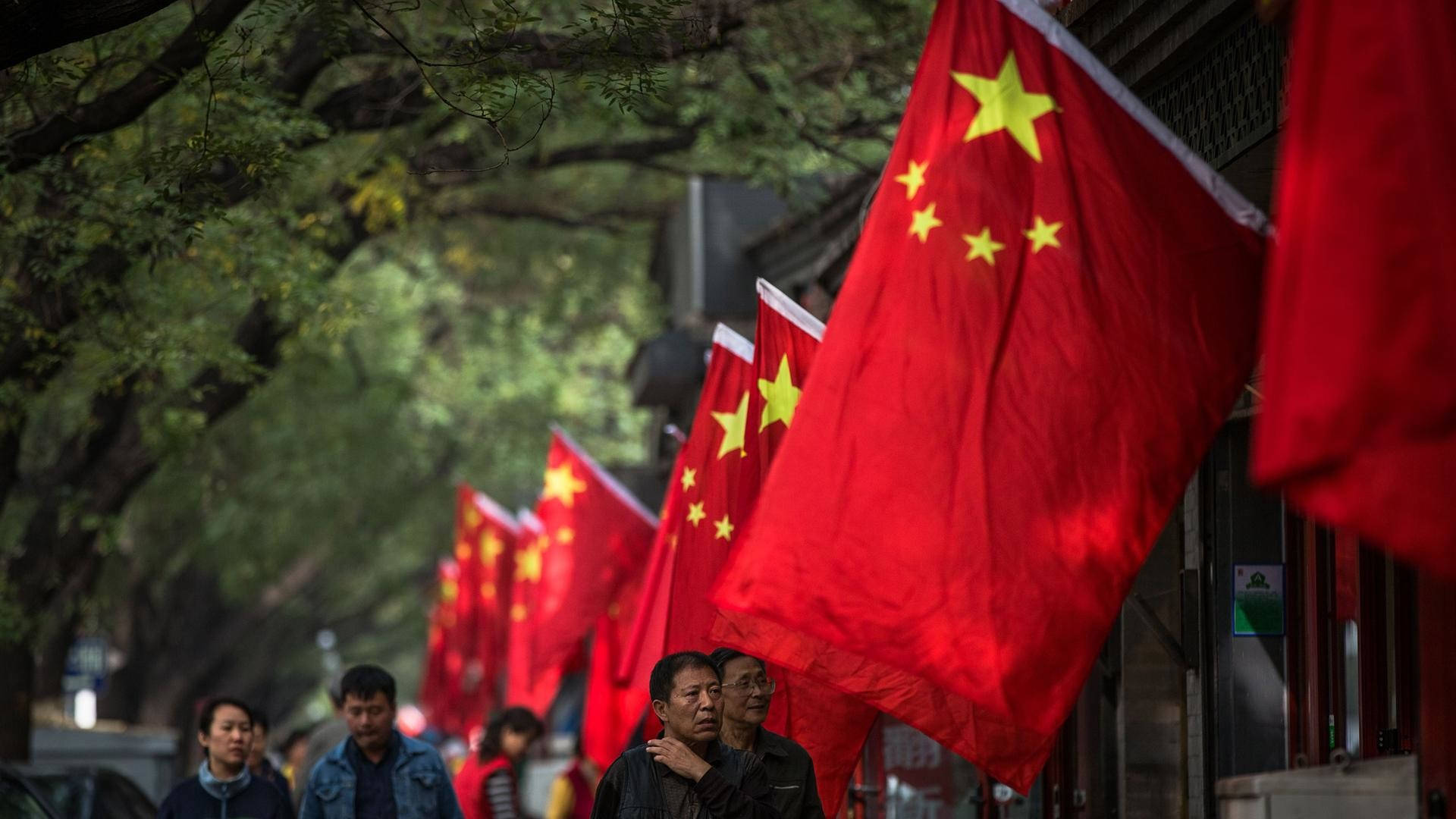中國國旗街景 壁紙 — 紅色中國國旗帶有黃色星星排列在一條樹蔭街道上，行人於白天走過。