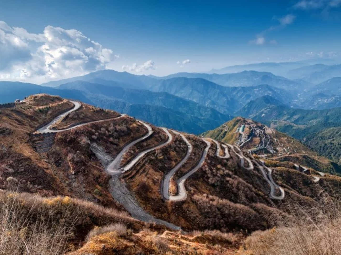 中國山間蜿蜒的道路 壁紙 — 一條蜿蜒的山路在明亮的藍天白雲下蜿蜒穿過秋日山丘。