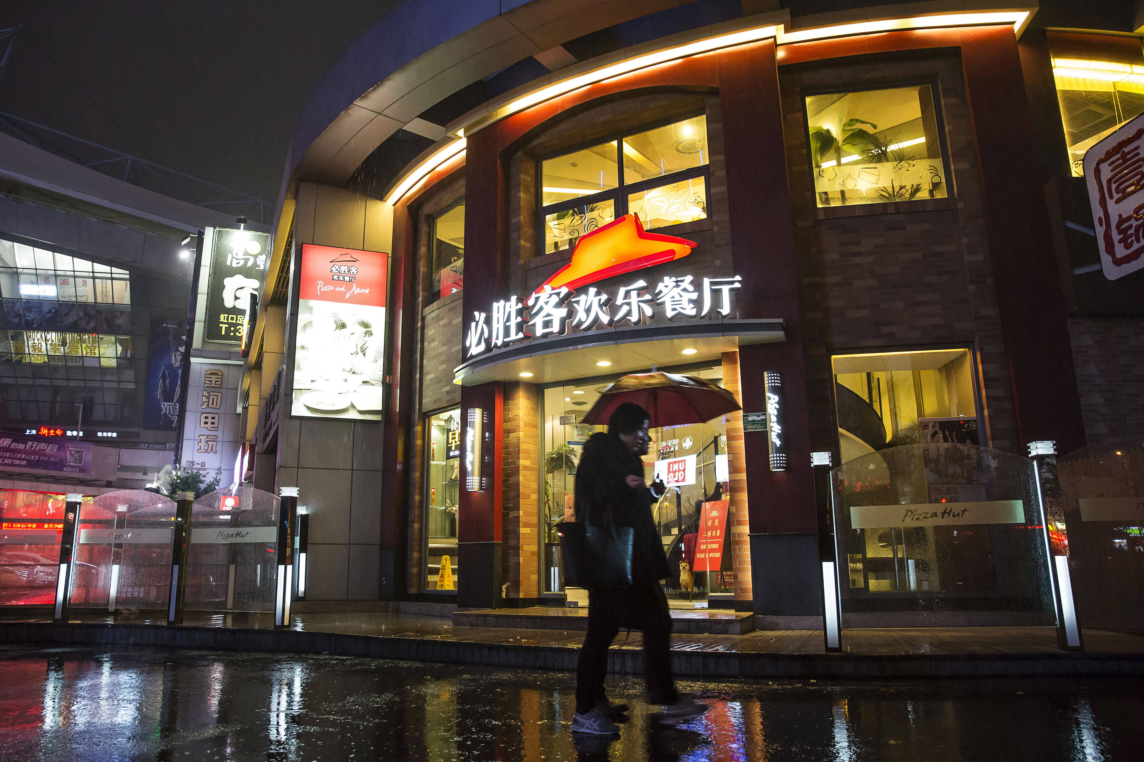 中國必勝客餐廳 壁紙 — 一個拿着雨傘的人夜晚在濕漉漉的、反射着霓虹燈光的城市街道上走過燈火通明的必勝客。