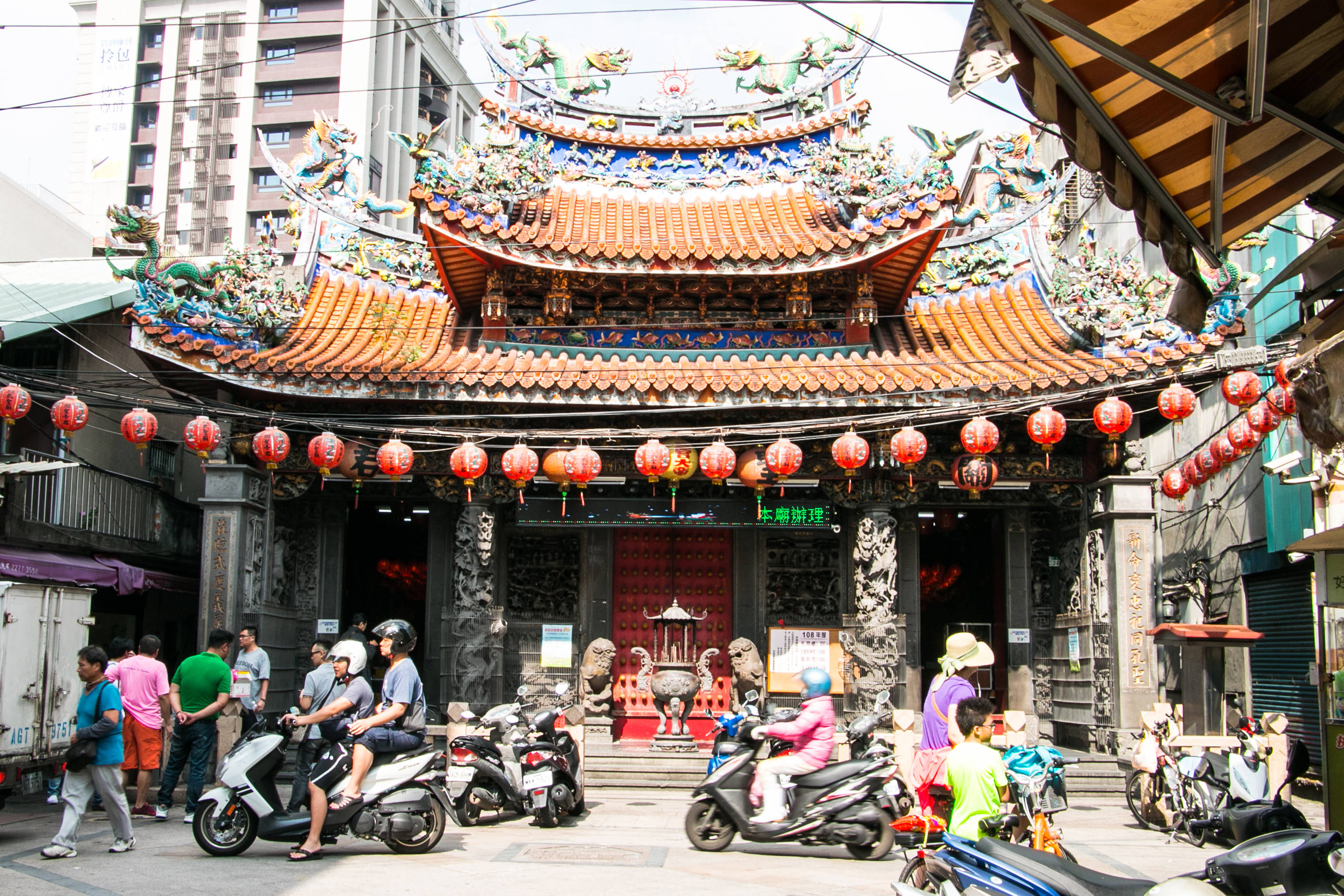 中國新年慶祝活動 壁紙 — 一座充滿活力的傳統寺廟，擁有華麗的瓦頂和紅燈籠，矗立在擠滿電單車和行人的繁華城市街道中。