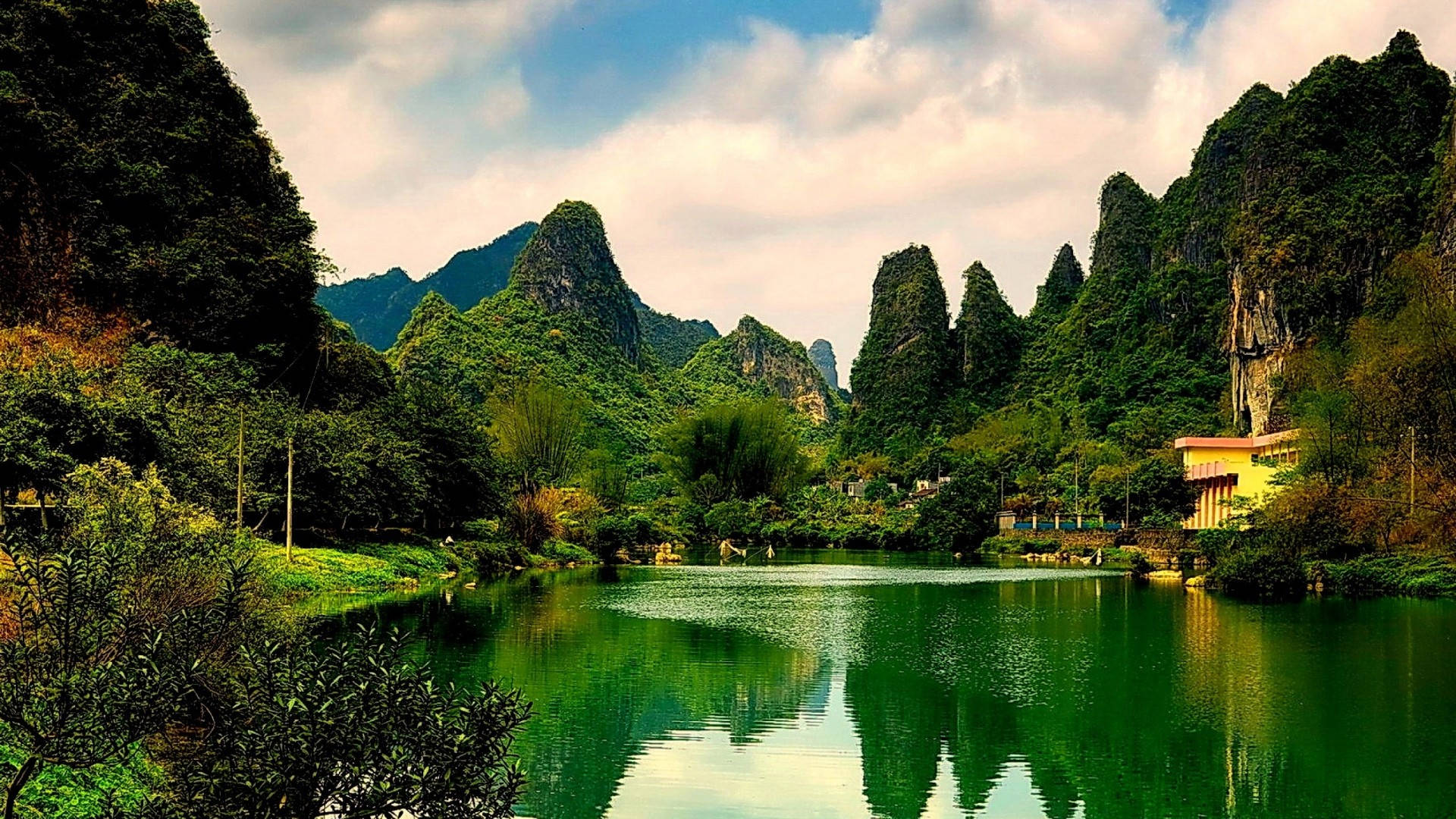 中國桂林石灰岩湖自然風景 壁紙 — 寧靜的湖面映照著鬱鬱蔥蔥的綠色喀斯特山脈，天空多雲，岸邊有一座小建築。