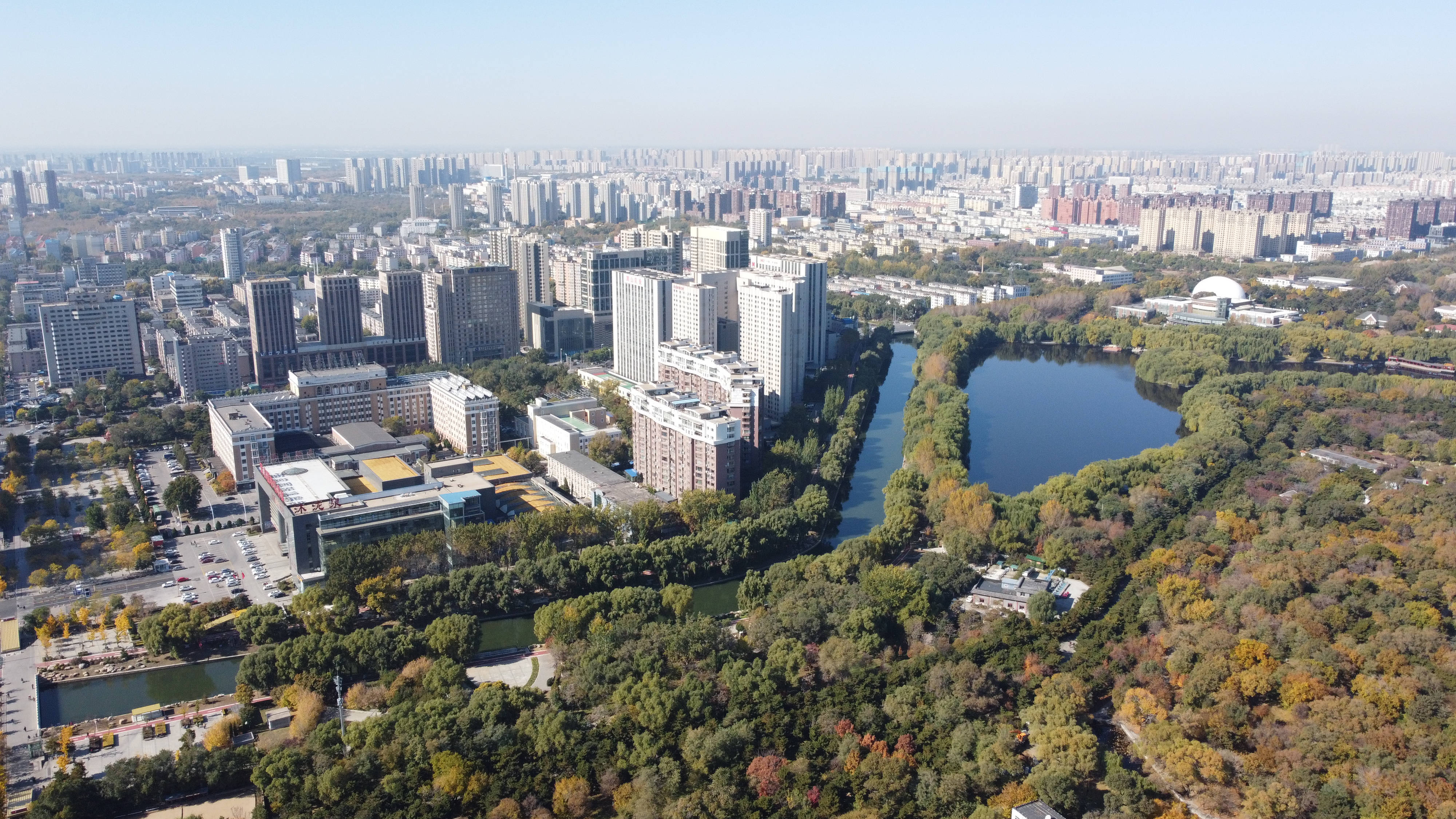 中國瀋陽城市 壁紙 — 從高空俯瞰廣闊的城市景觀，現代高層建築坐落在秋色樹木之中，環繞著蜿蜒的河流和一個大湖。