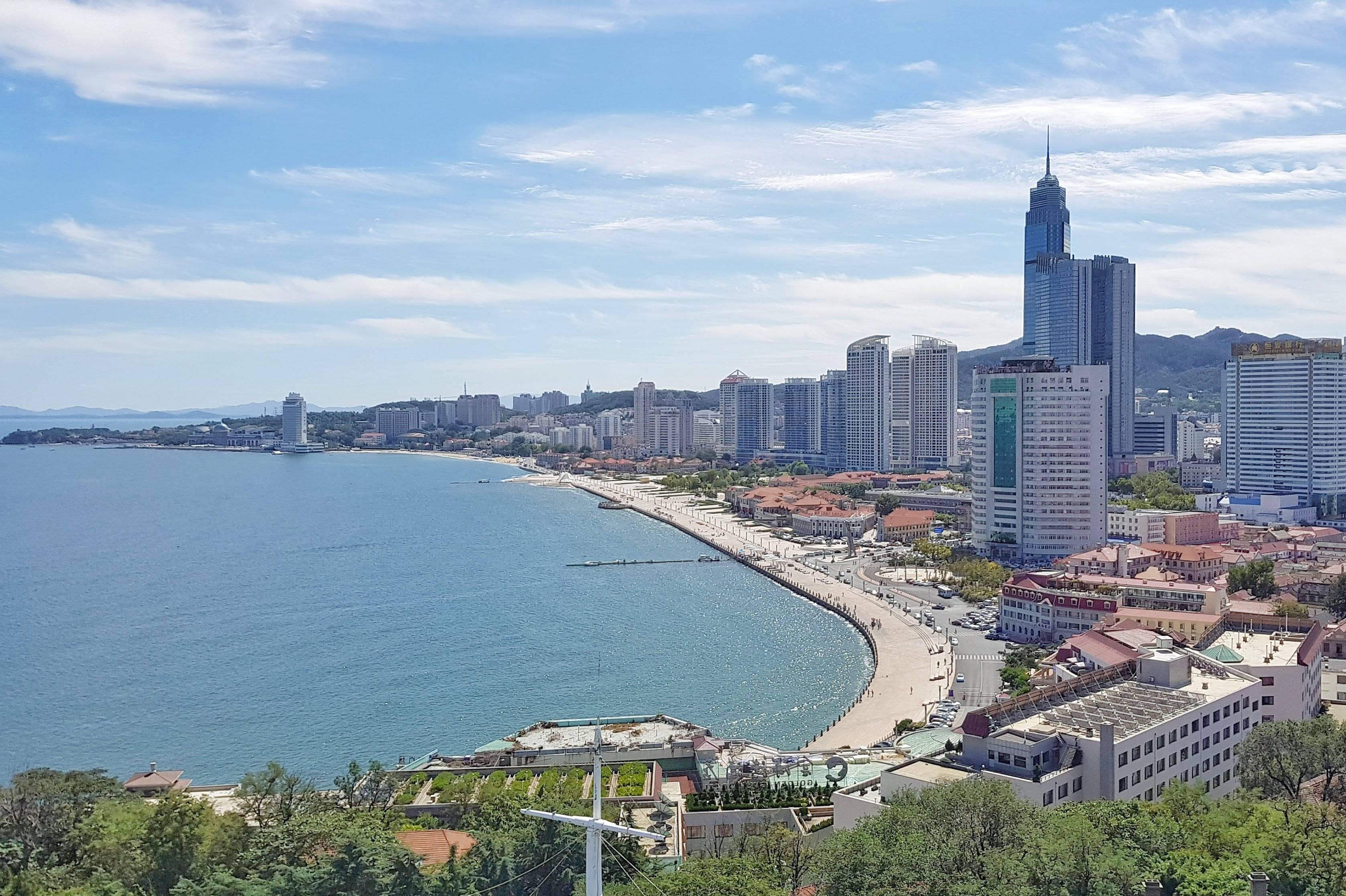 中國煙台城市 壁紙 — 海岸城市的全景視圖，現代摩天大樓沿著彎曲的海灘排列，在明亮的藍天白雲下。