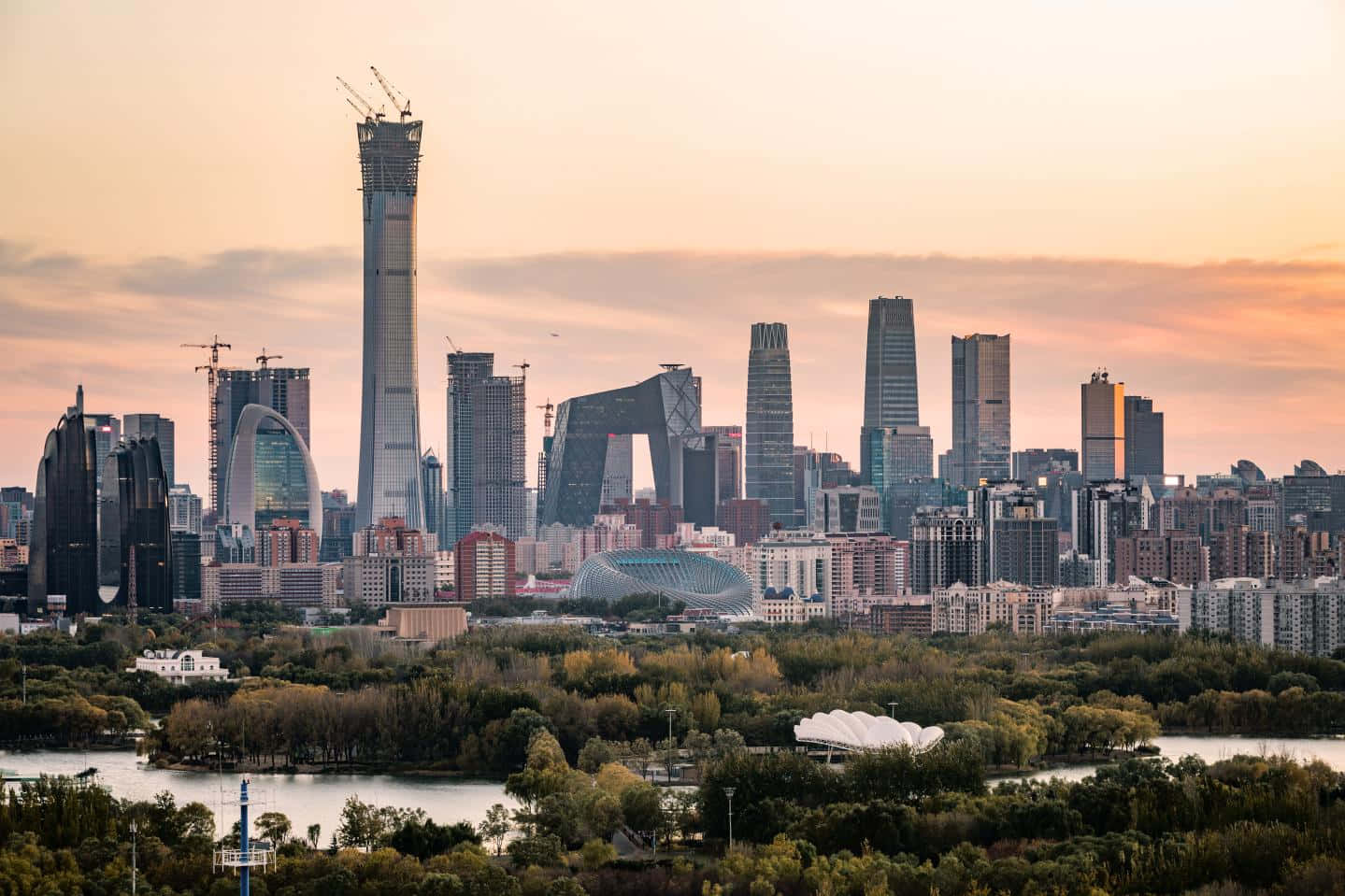 中國的輝煌 壁紙 — 日落時分現代城市天際線的全景視圖，高樓大廈和建築起重機聳立在河流和綠色公園之上。