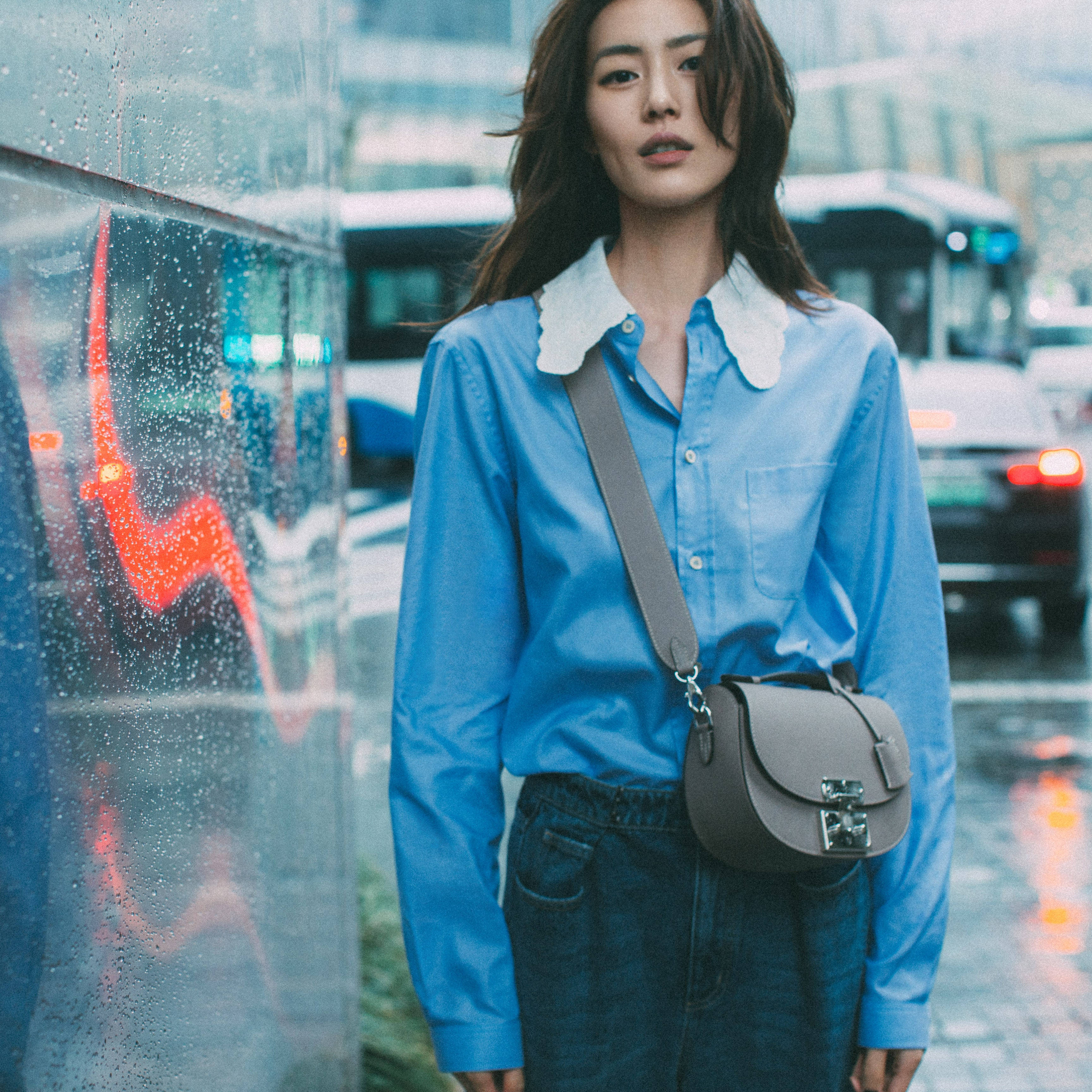 中國超模 Liu Wen 搭配 Moynat 手袋 壁紙 — 一位身穿藍色白領襯衫的女性，在夜晚的城市街道上站在濺滿雨水的窗邊。