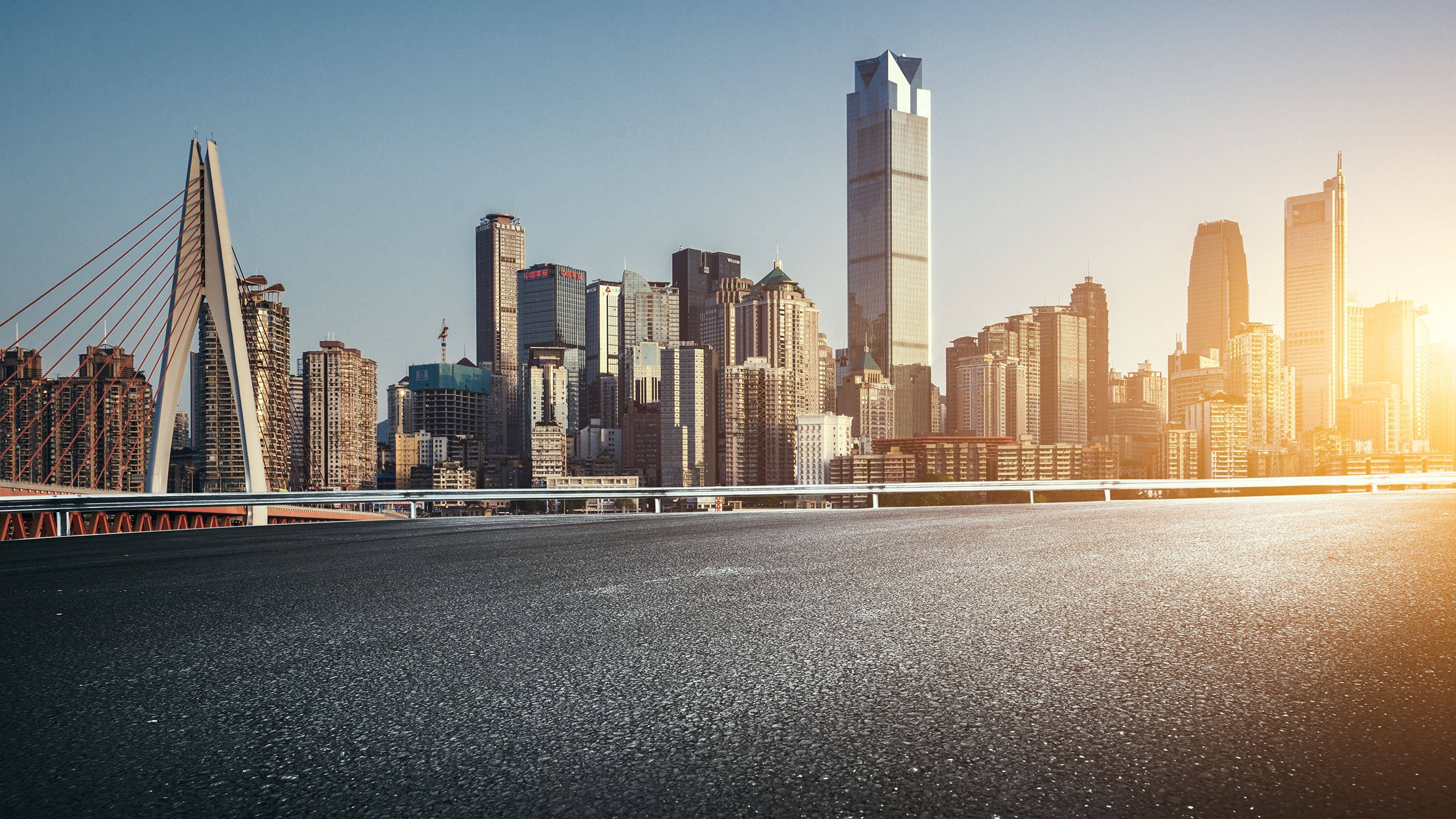 中國重慶街景視角 壁紙 — 夕陽以金色光芒沐浴現代城市景觀，一座橋樑橫跨寬闊的河流，摩天大樓在後方聳立。