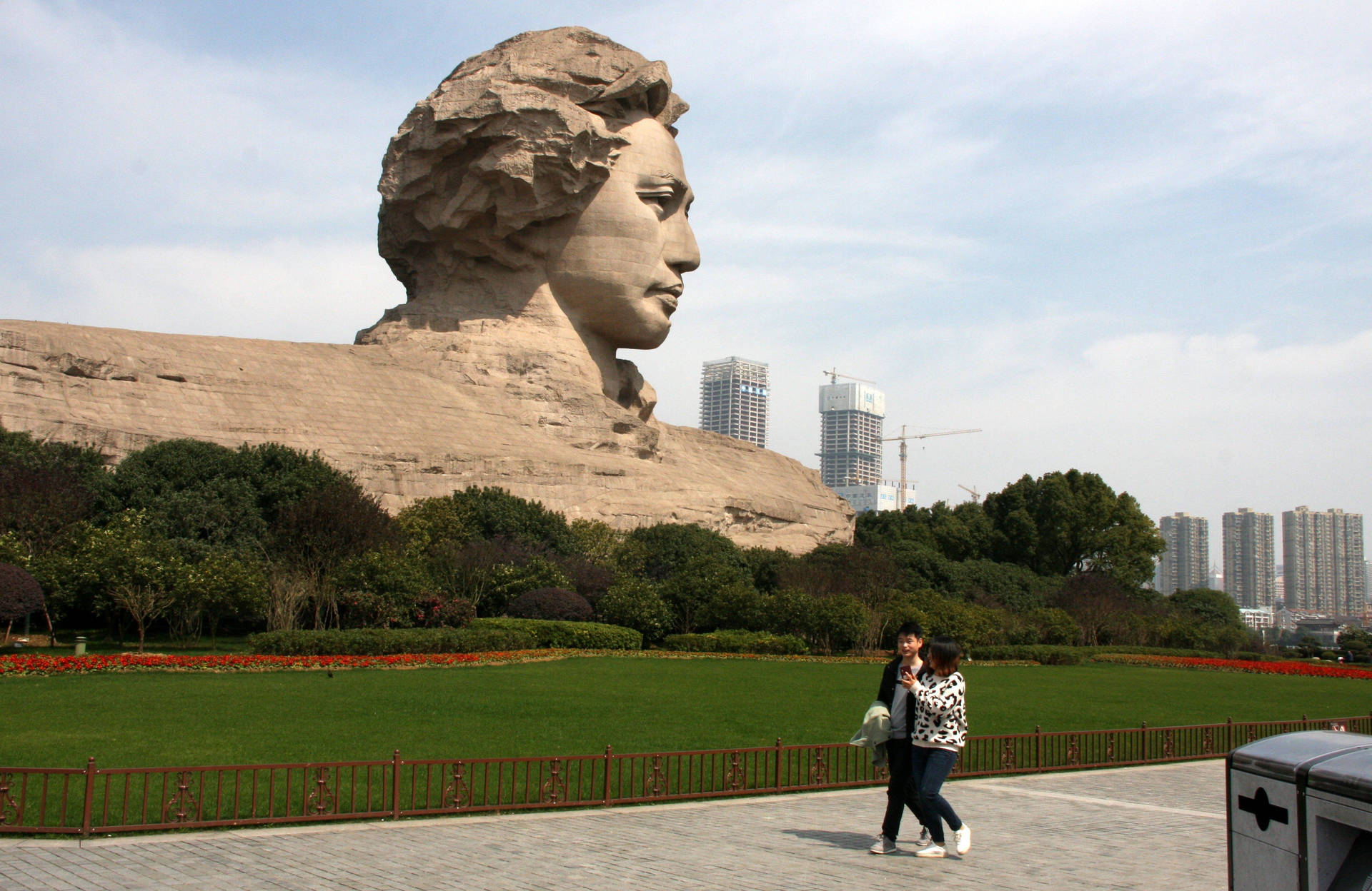 中國長沙城市 壁紙 — 一個巨大的女性頭部石雕俯瞰著公園，公園裡有兩個人走在鋪砌的小徑上。