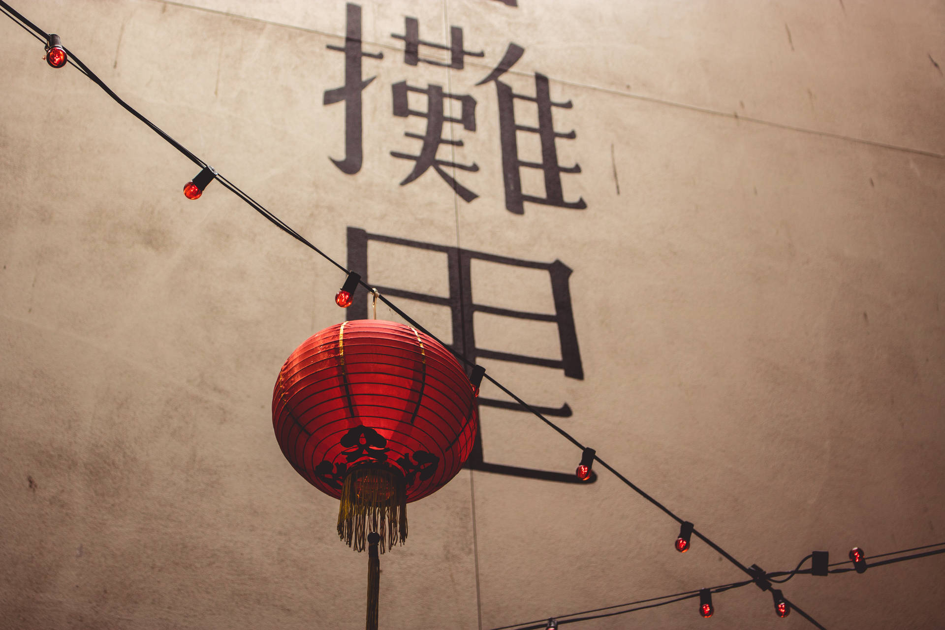 紅色燈籠帶流蘇掛在帶有中文字和燈串的牆前。
