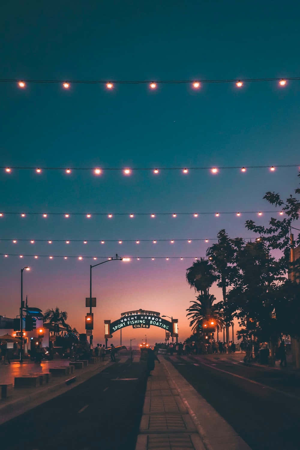 Spring Lights 桌布 壁紙 — 串燈在黃昏時分溫暖地照亮了熱鬧的海濱長廊，伴隨著棕櫚樹和發光的拱門。