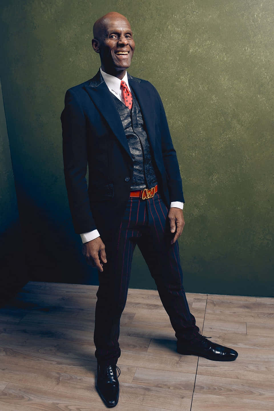一位身穿時尚西裝的快樂男士，站在鋪有木地板的綠色牆壁工作室中。