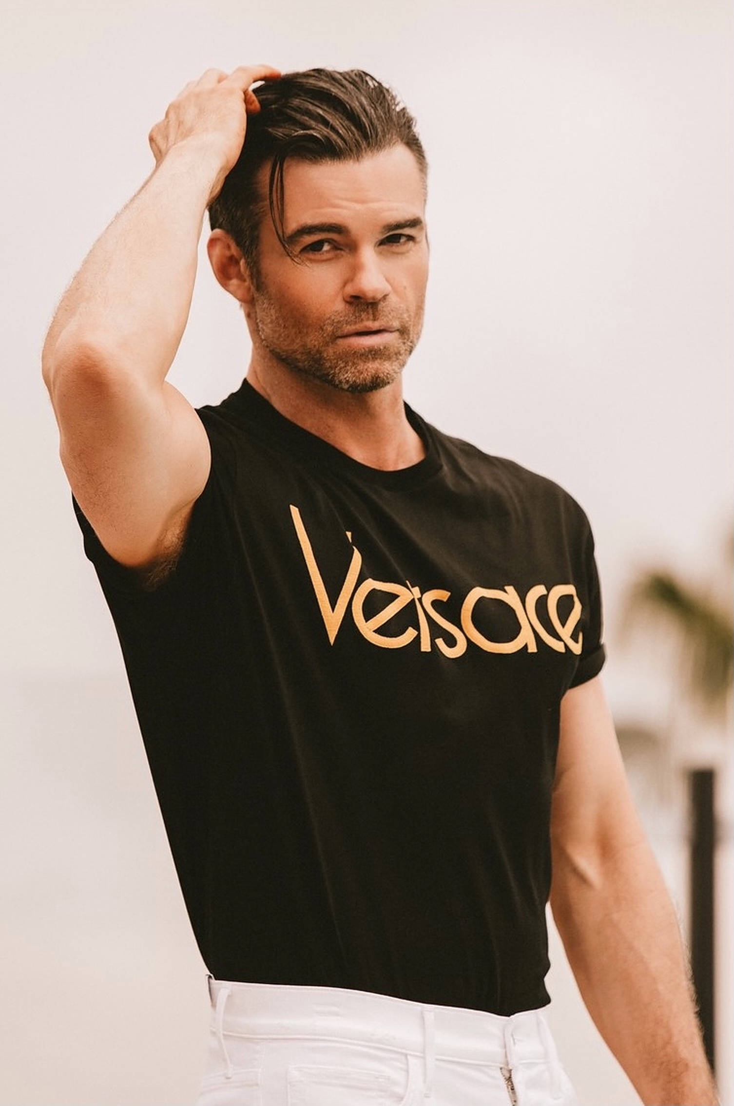 丹尼爾·吉利斯 Versace 壁紙 — 一名身穿黑色Versace T恤和白色褲子的男子，在柔和模糊的背景前，用手梳理他造型過的頭髮。
