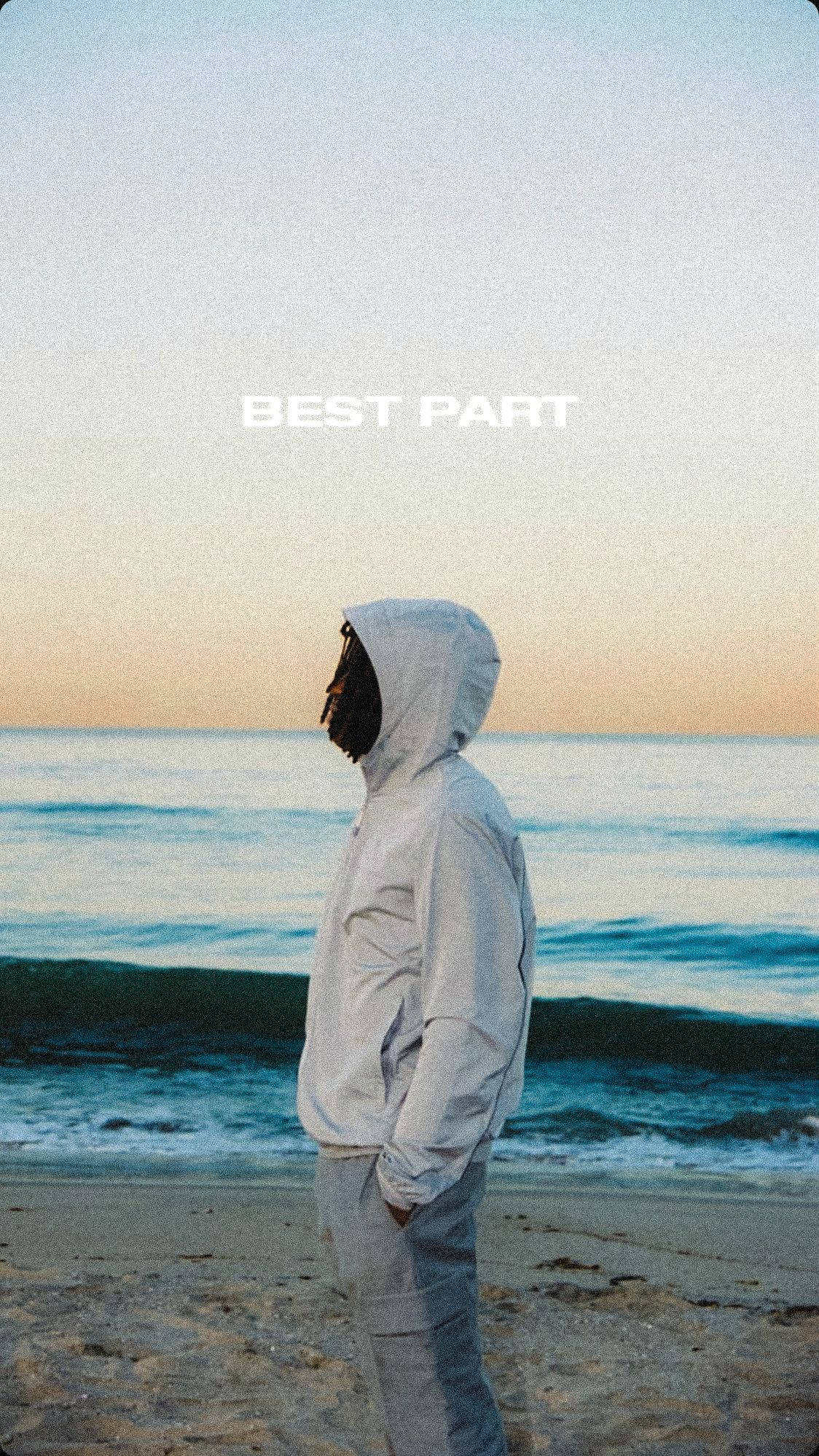 丹尼爾·西薩爾《Best Part》 壁紙 — 一個穿著白色連帽衫的人獨自站在日落時分的沙灘上，凝視著波濤洶湧的大海。