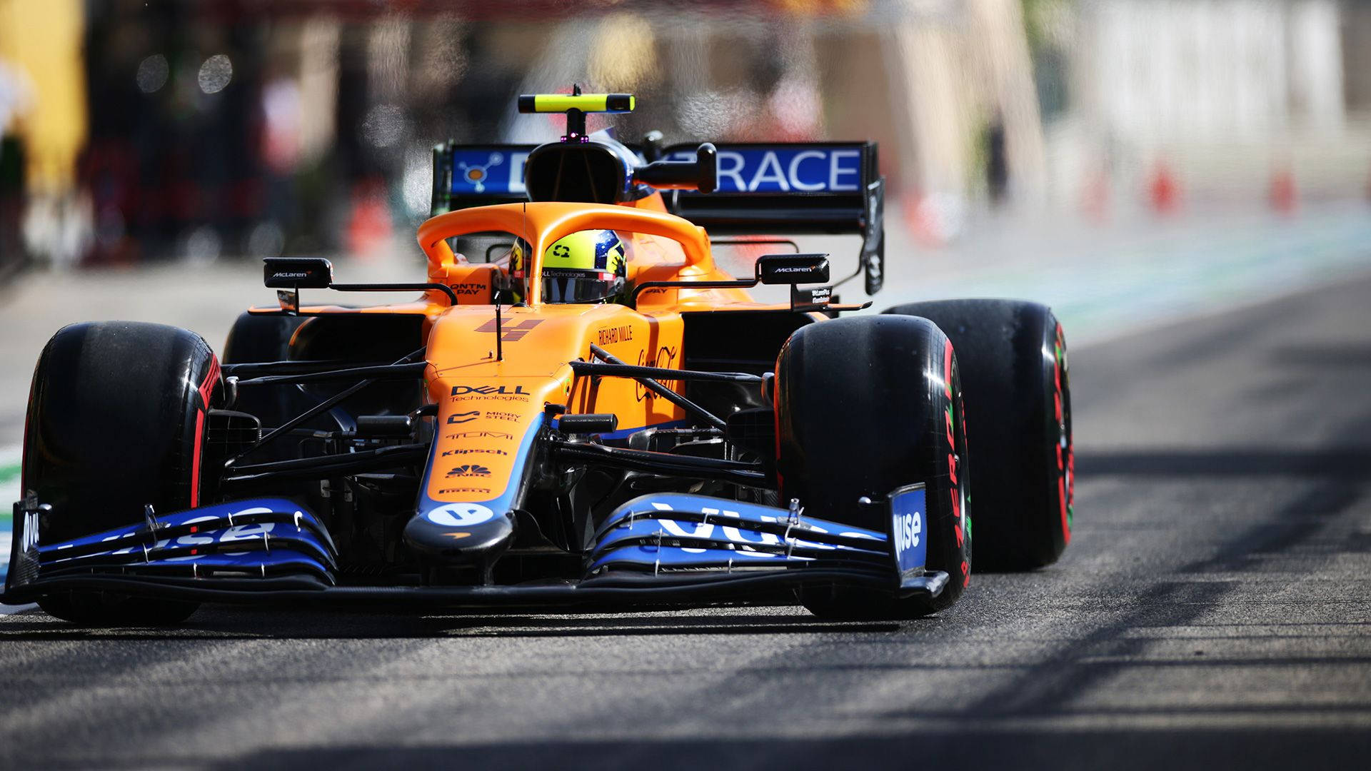 丹尼爾·里卡多F1賽車在賽道上 壁紙 — 一輛充滿活力的橙藍色一級方程式賽車在陽光明媚的賽道上疾馳，背景錐體模糊。
