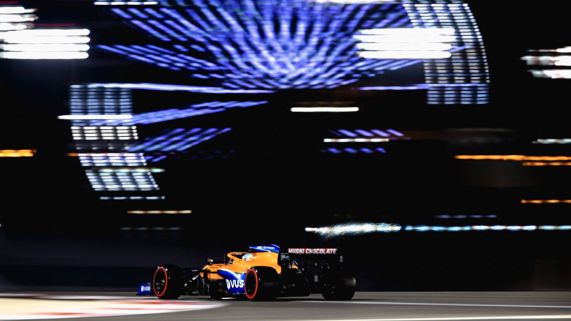 丹尼爾·里卡多F1賽車對比大型廣告牌 壁紙 — 一輛充滿活力的橙色和藍色一級方程式賽車在夜間於賽道上奔馳，伴隨著耀眼的藍色和白色光條。