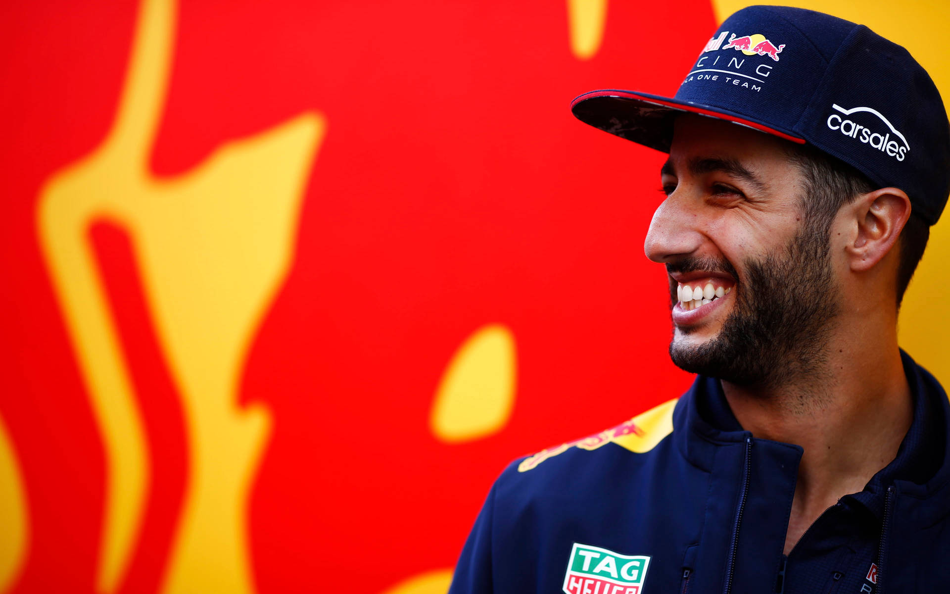 丹尼爾·里卡多側臉微笑 壁紙 — 一名面帶微笑的男子身穿Red Bull Racing車隊外套和帽子，站在充滿活力的紅黃抽象背景前。