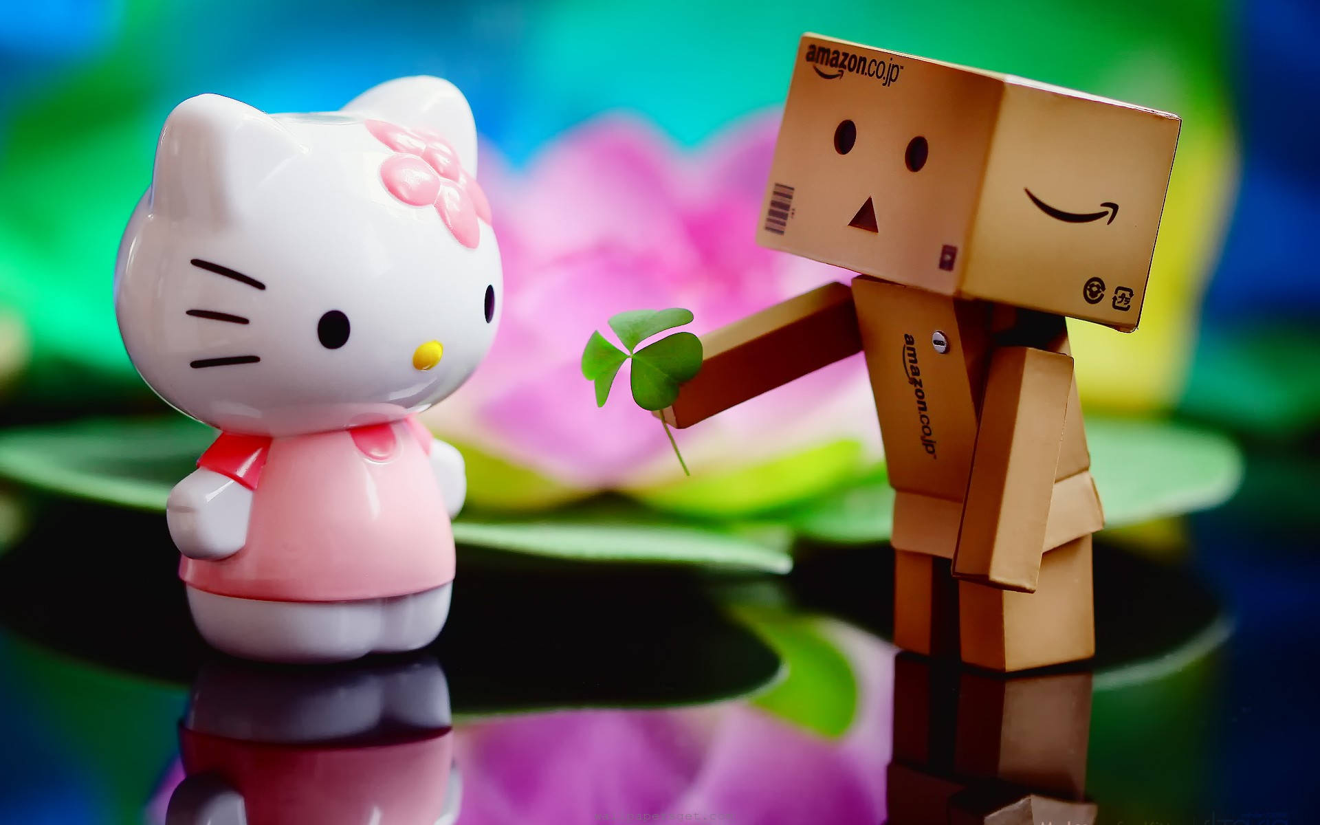 丹波與Hello Kitty桌面擺設 壁紙 — Hello Kitty 公仔喺色彩繽紛模糊嘅花卉背景前，從一個微笑嘅紙板 Danbo 機械人度收下四葉草。