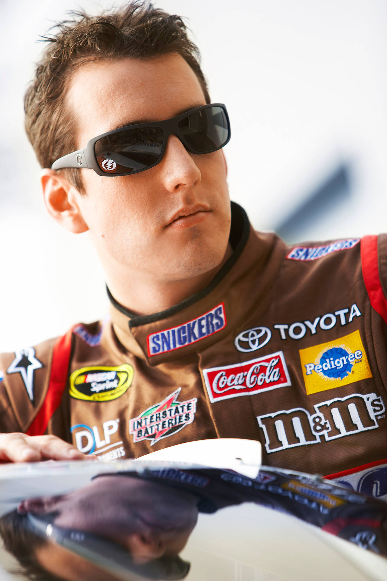 主宰NASCAR的冠軍——凱爾·布希 壁紙 — 一位專注的NASCAR車手穿著佈滿贊助商標誌的棕色賽車服和太陽眼鏡，俯身靠在汽車引擎蓋上。