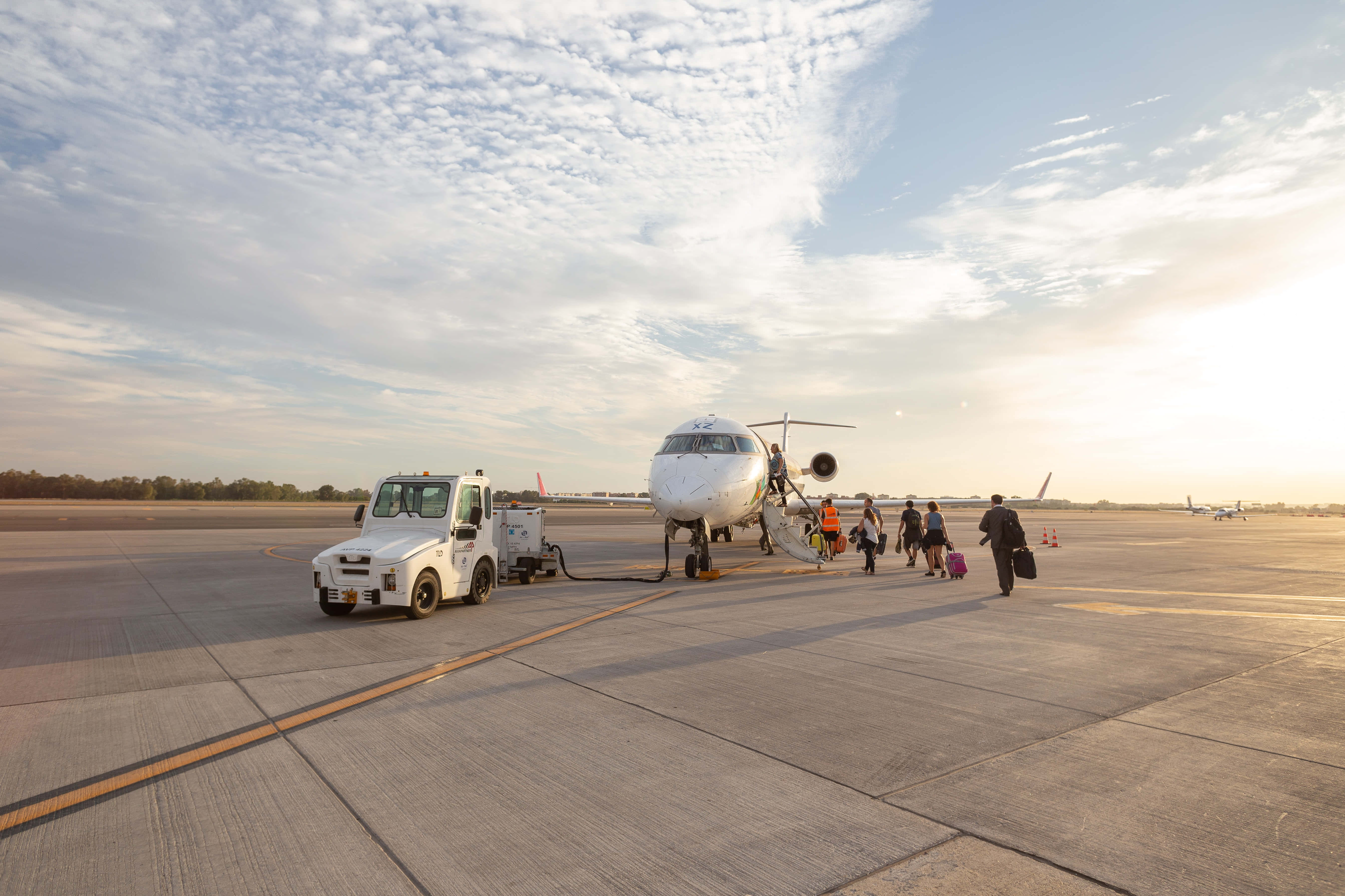Airport 桌布 壁紙 — 乘客在陽光明媚的停機坪上走向一架小型飛機，附近有一輛地面車輛停在部分多雲的天空下。