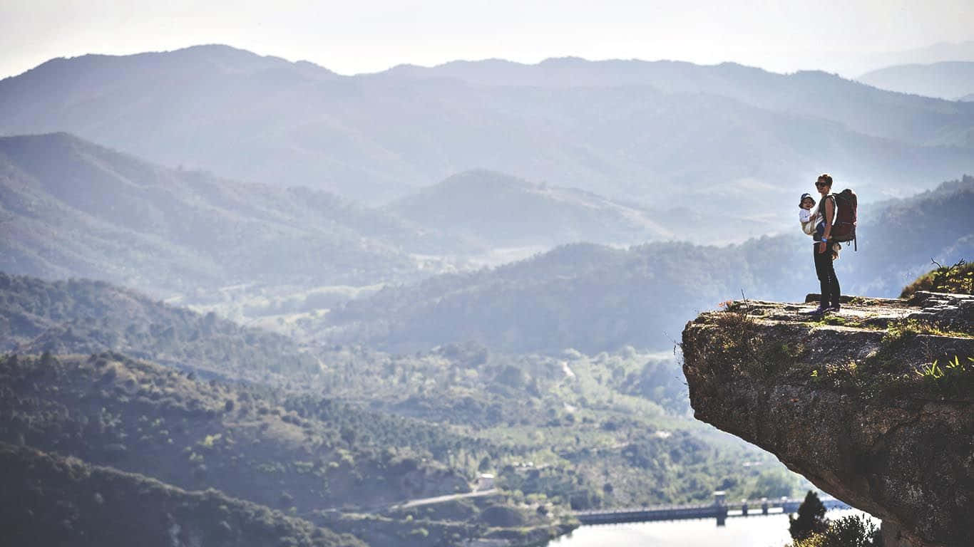 乘風征服山巒 壁紙 — 一位孤獨的遠足者揹著嬰兒在揹架上，站在岩石峭壁頂端，俯瞰著沐浴在柔和晨光中的廣闊山谷。