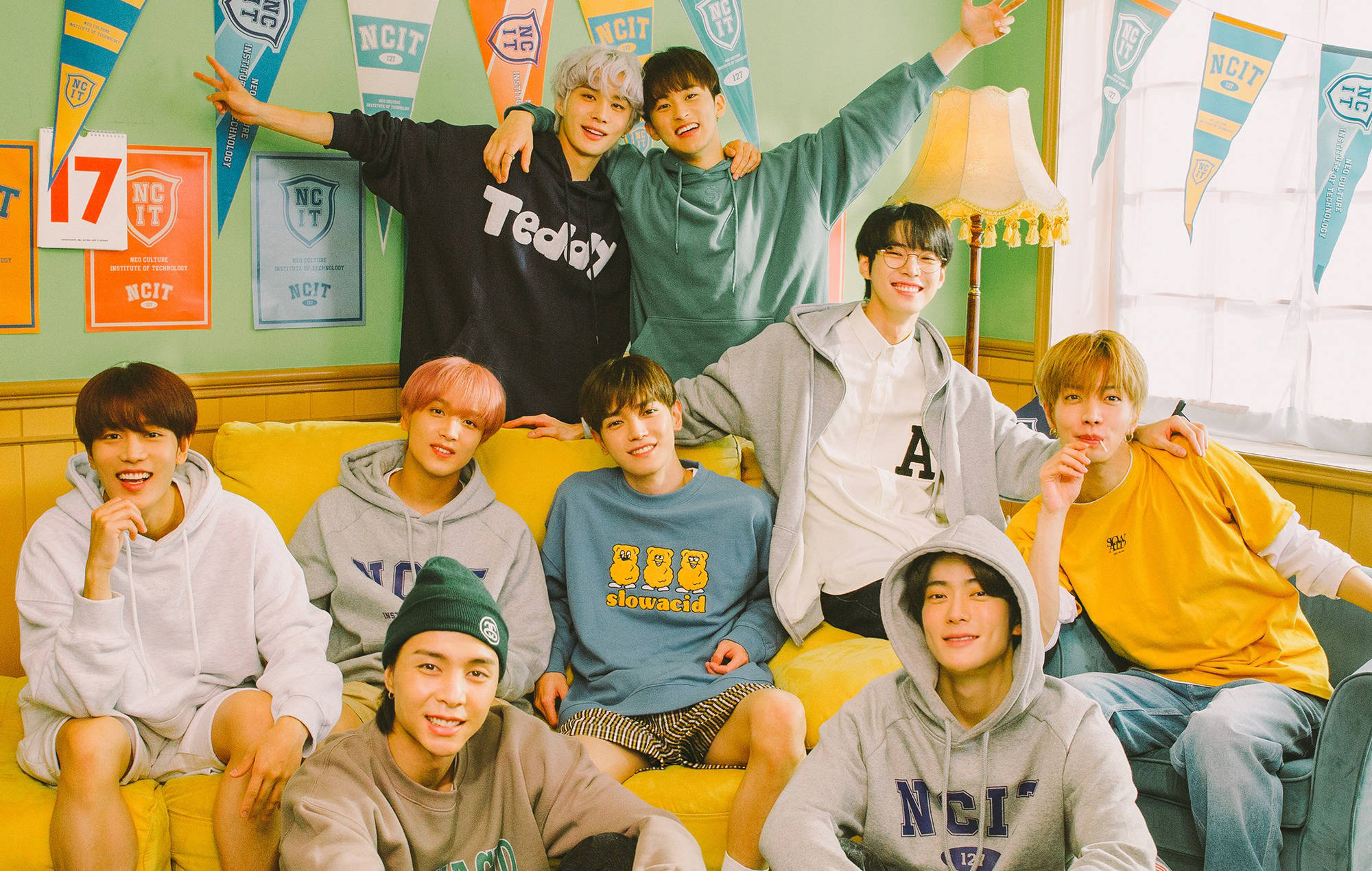 NCT 127 NCIT House 壁紙 — 九名年輕男子穿著休閒連帽衫和T恤，在裝飾有NCIT橫幅的明亮房間裡的黃色沙發上愉快地擺姿勢。