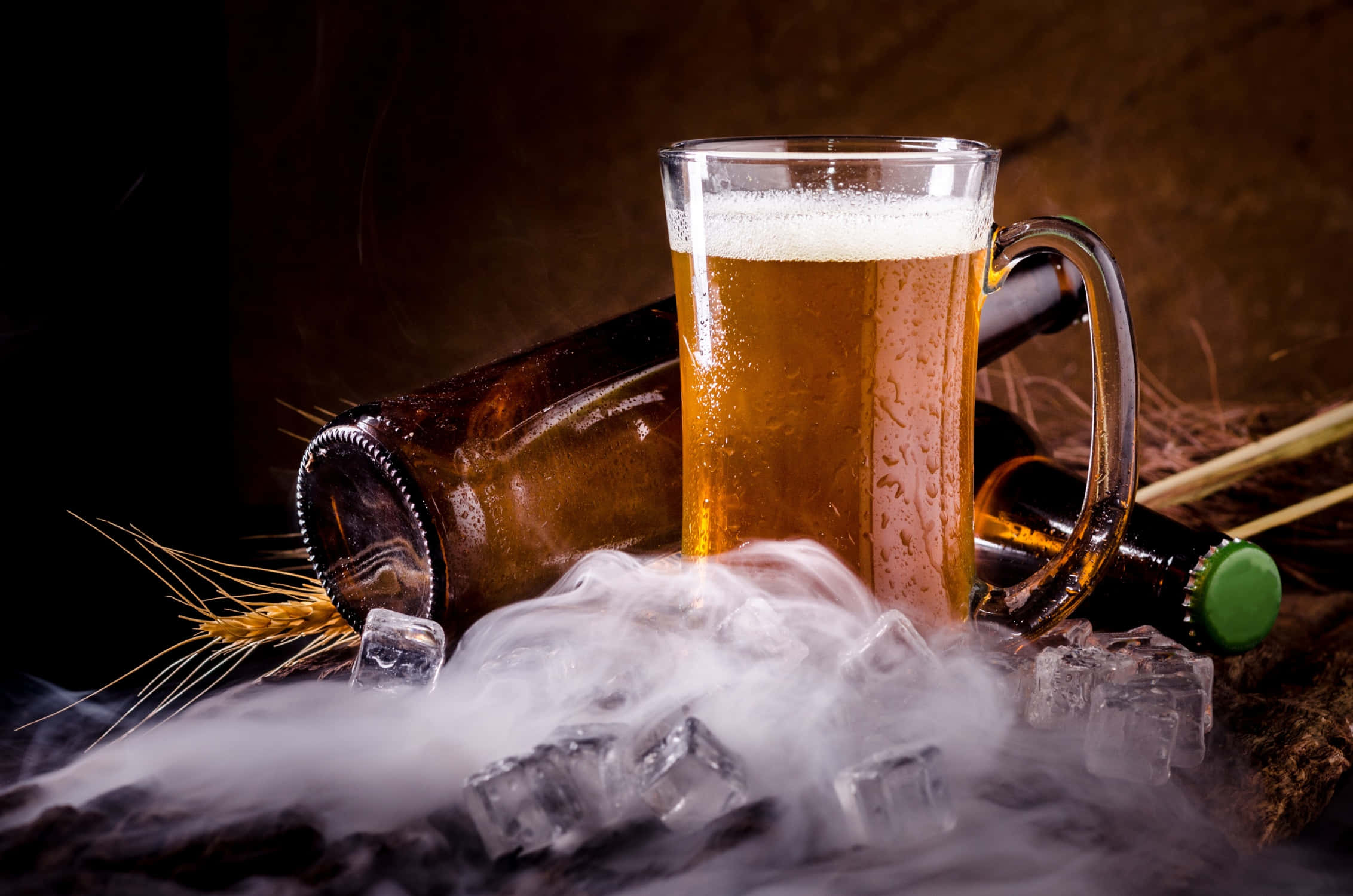 「乾杯！沒有比夏日冰啤酒更棒的了！」 壁紙 — 一杯冰凍琥珀色啤酒連泡沫放在乾冰煙霧上，旁邊有小麥穗和空瓶，在陰暗的氛圍中。