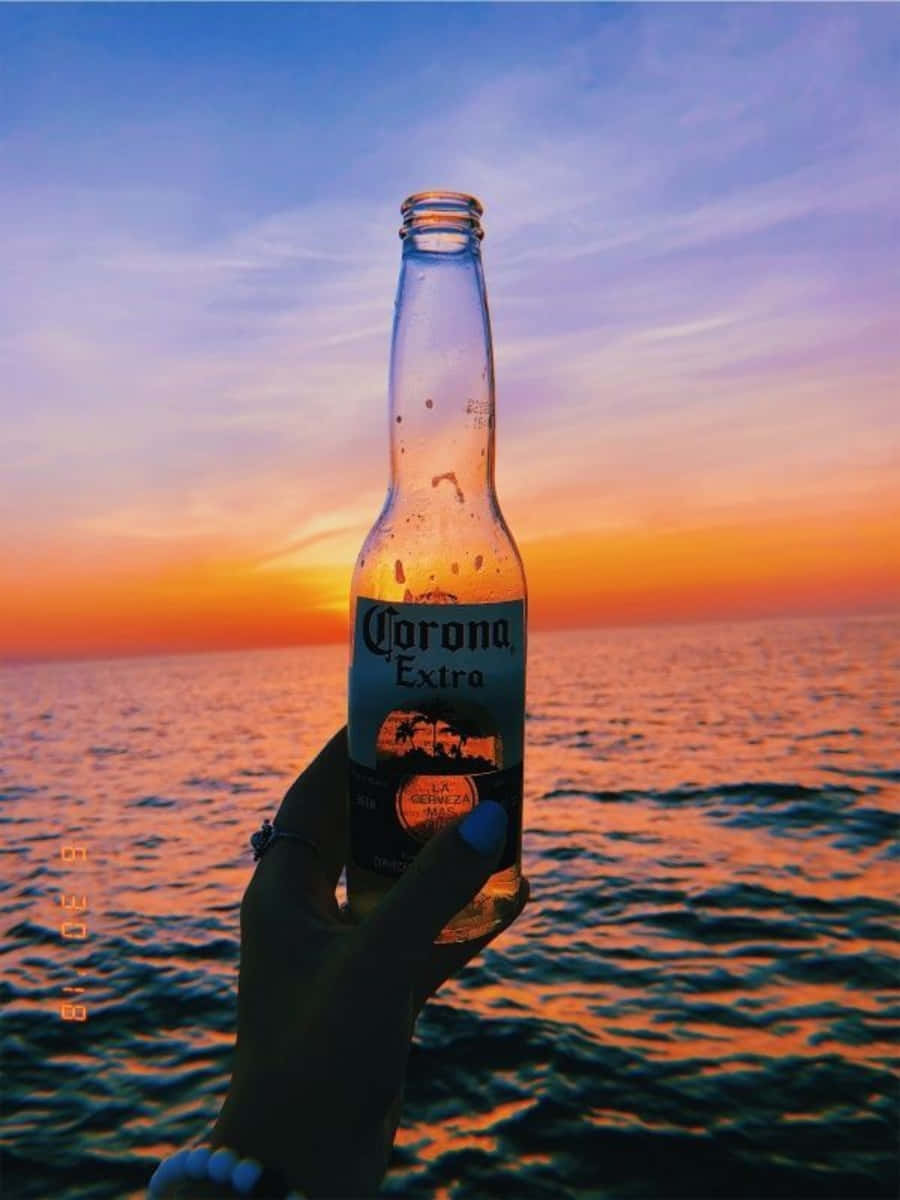 「乾杯！炎熱夏日後的清爽啤酒」 壁紙 — 一隻手拿著冰凍的Corona Extra啤酒瓶，背景是海洋上充滿活力的日落，天空呈現橙色和紫色。