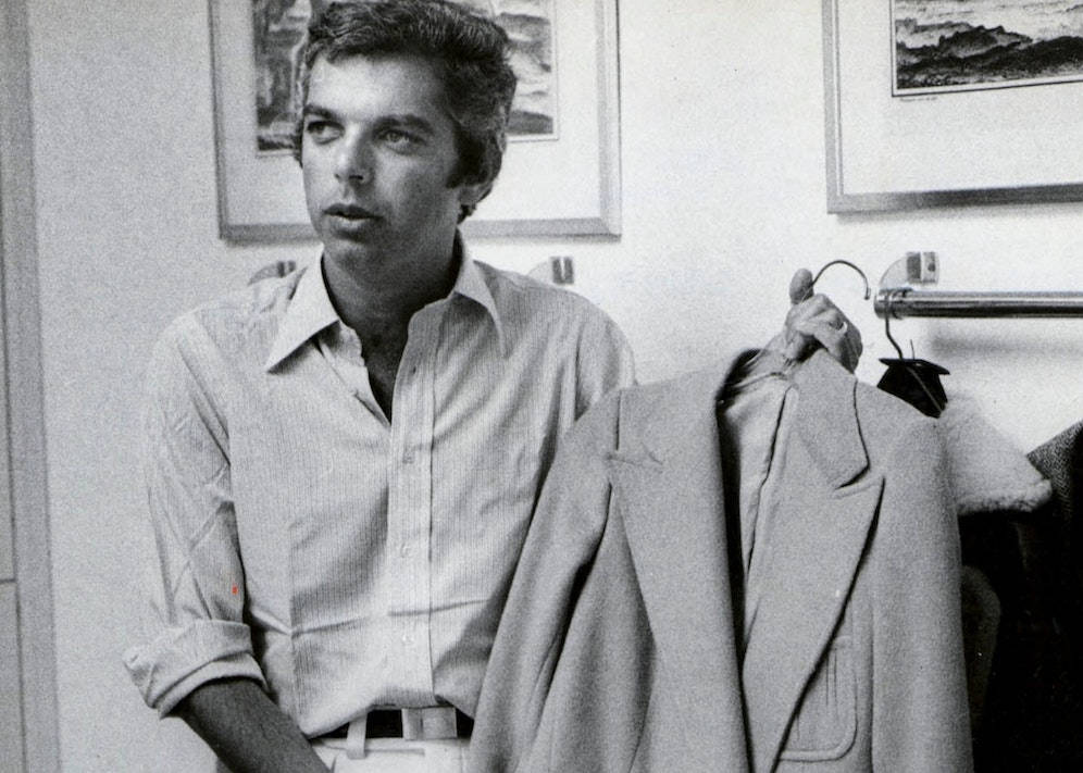 二十多歲的 Ralph Lauren 壁紙 — 一位穿著鈕扣襯衫的年輕男子，在柔和光線的房間裡，牆上掛著裱框藝術品，若有所思地凝視著，同時拿著一件西裝外套。