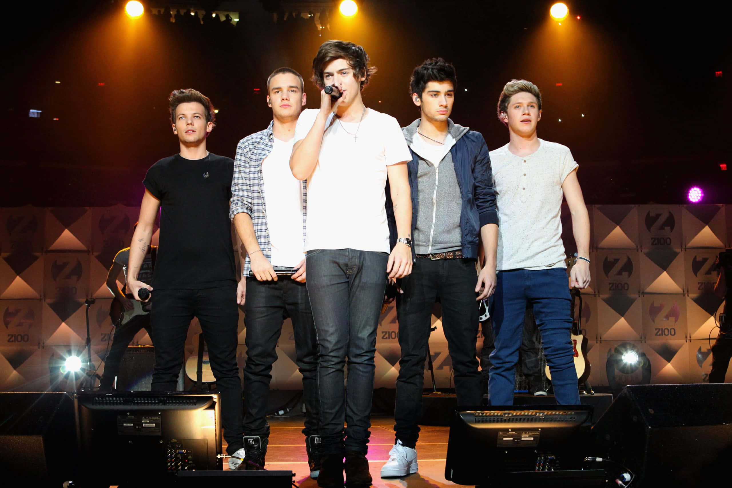 One Direction 桌布 壁紙 — 五位年輕男性樂隊成員在燈光舞台上表演，其中一人對著麥克風唱歌。